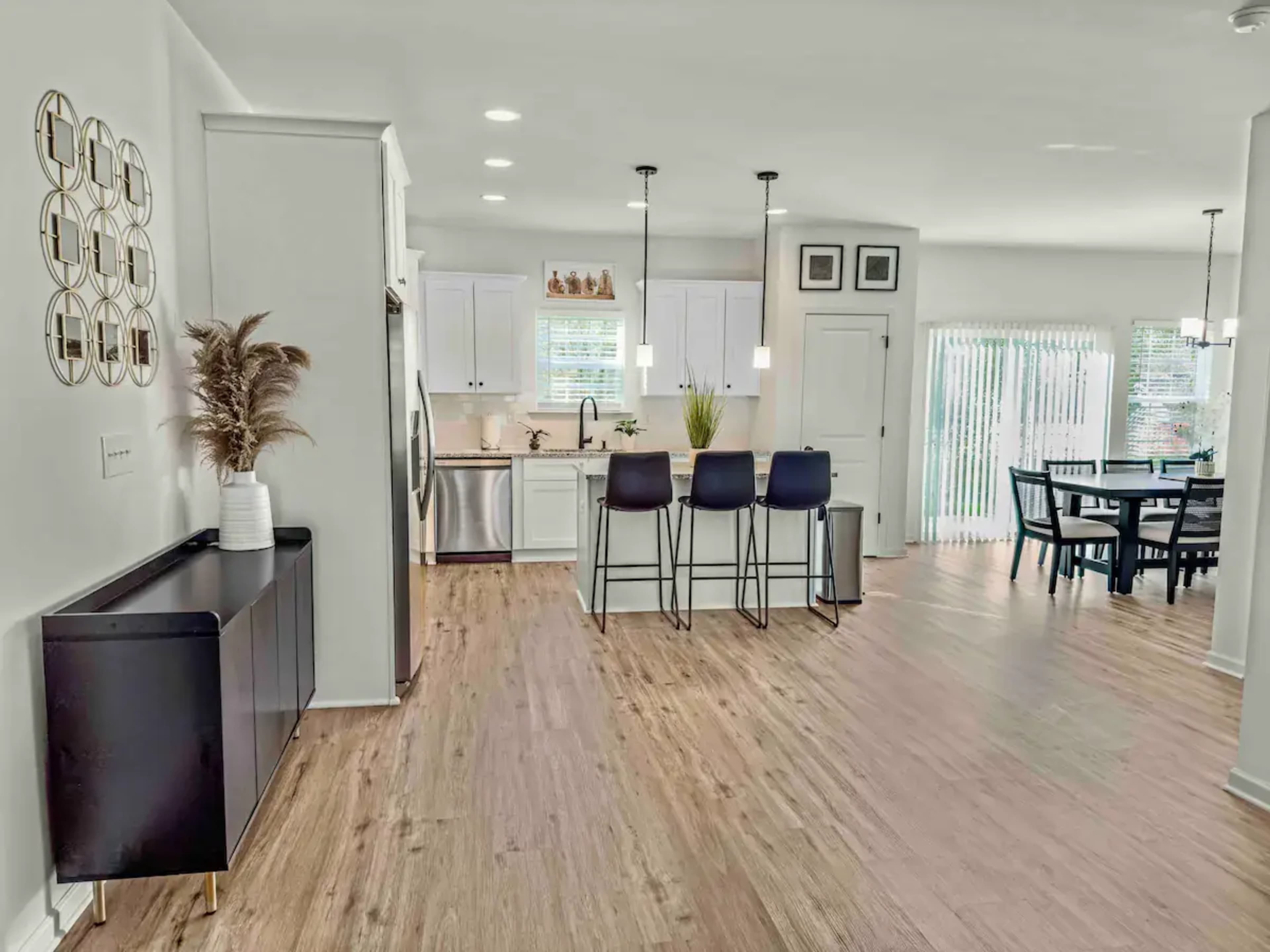 The image shows a modern open-concept kitchen and dining area with sleek cabinets, bar stools, and a dining table in a well-lit room.