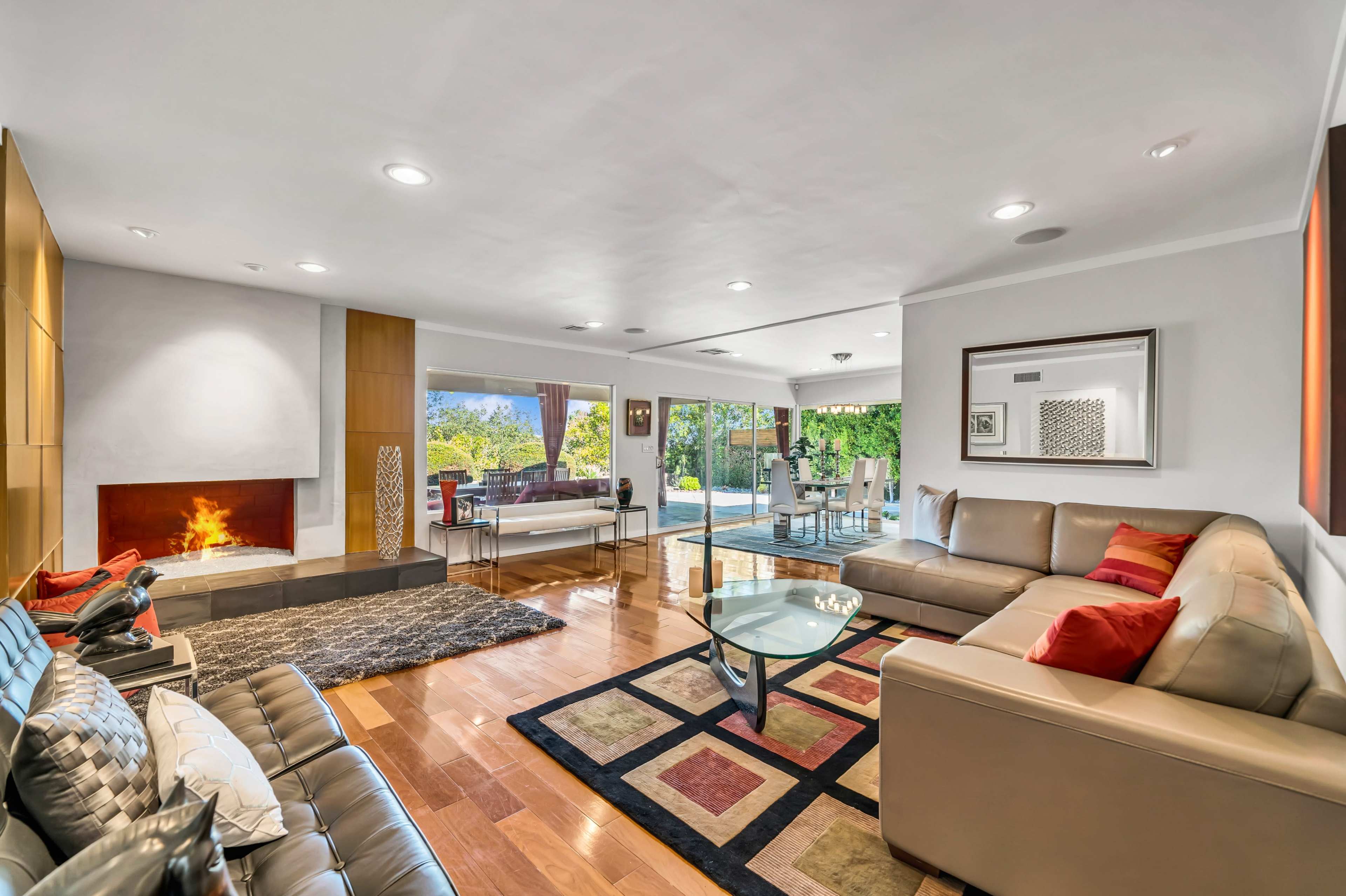 The living room features modern furnishings with a sectional sofa, a glass coffee table, and a fireplace, all arranged around a large window that provides a view of the outdoors.