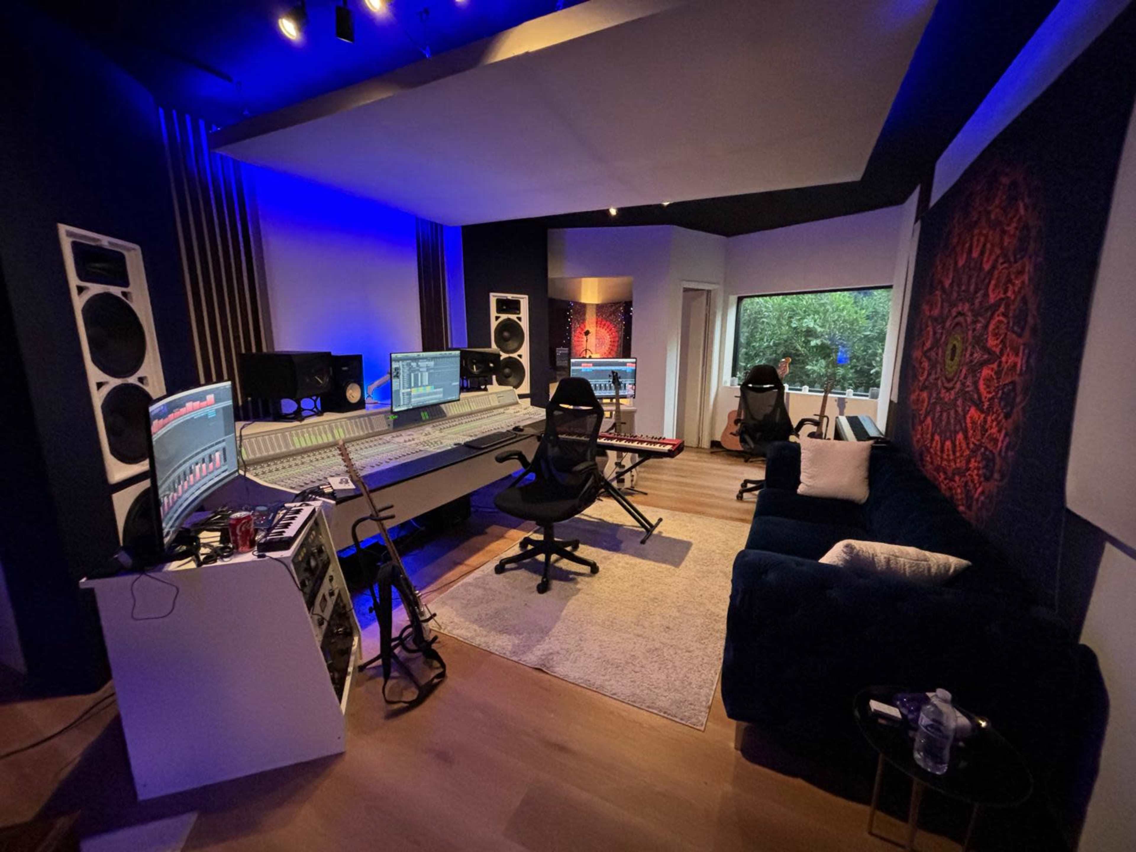 A music recording studio features a large mixing desk, speakers, and a couch, with ambient lighting and a window overlooking greenery.