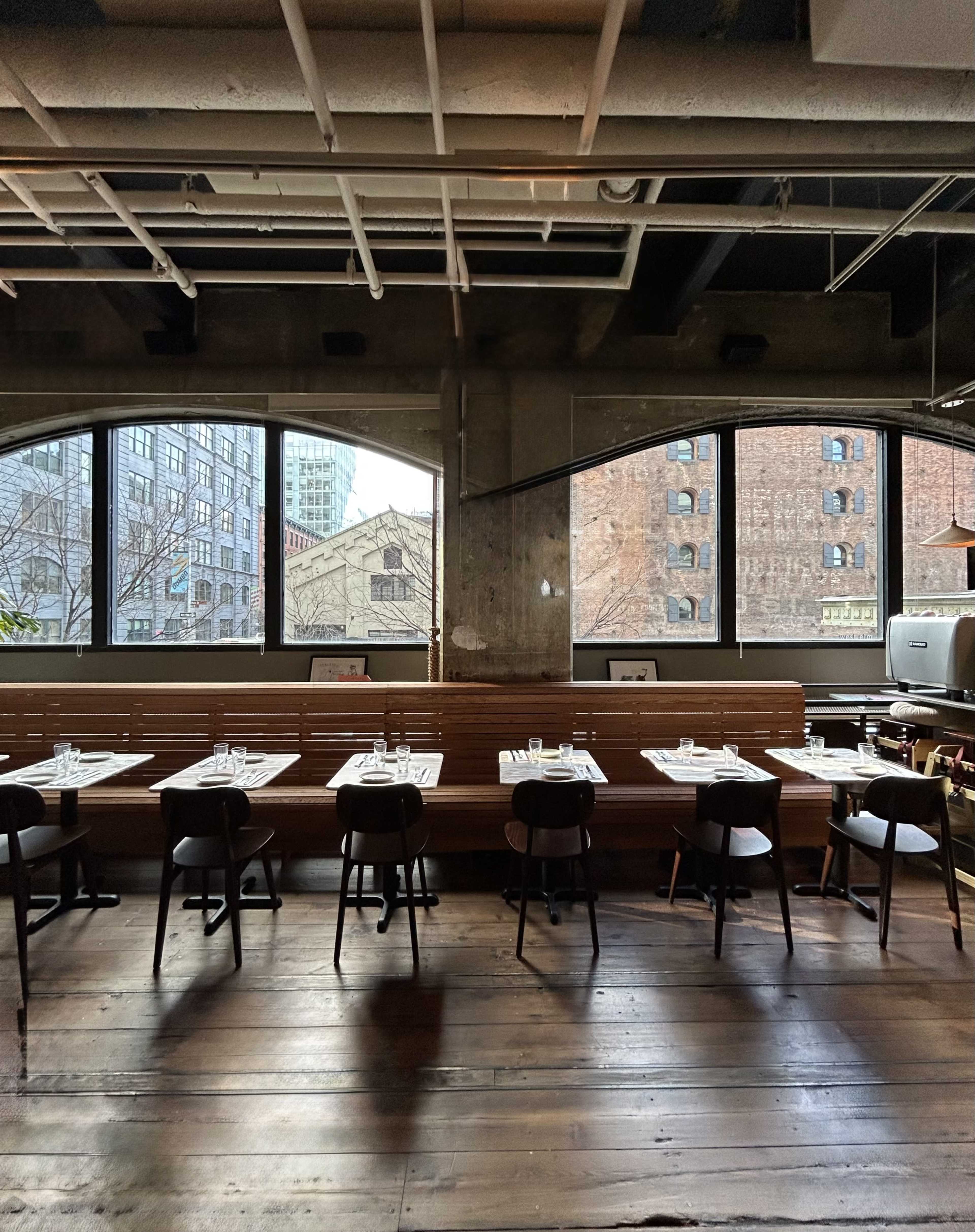 The image shows a modern restaurant interior featuring wooden tables set for dining and large windows revealing an urban view outside.