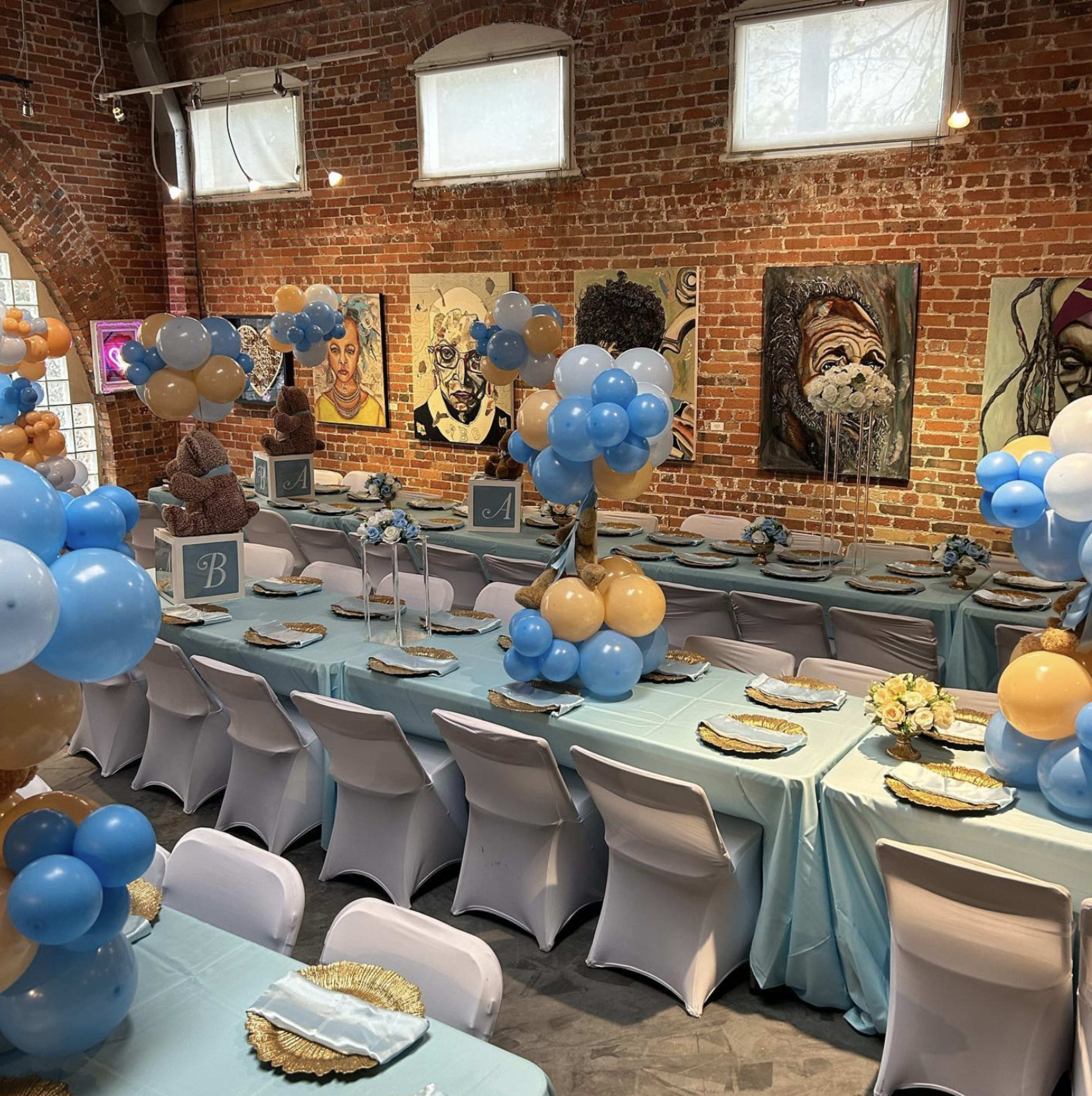 A decorated event space features tables set with blue and gold tablecloths, balloons, and floral centerpieces against a backdrop of brick walls and artwork.