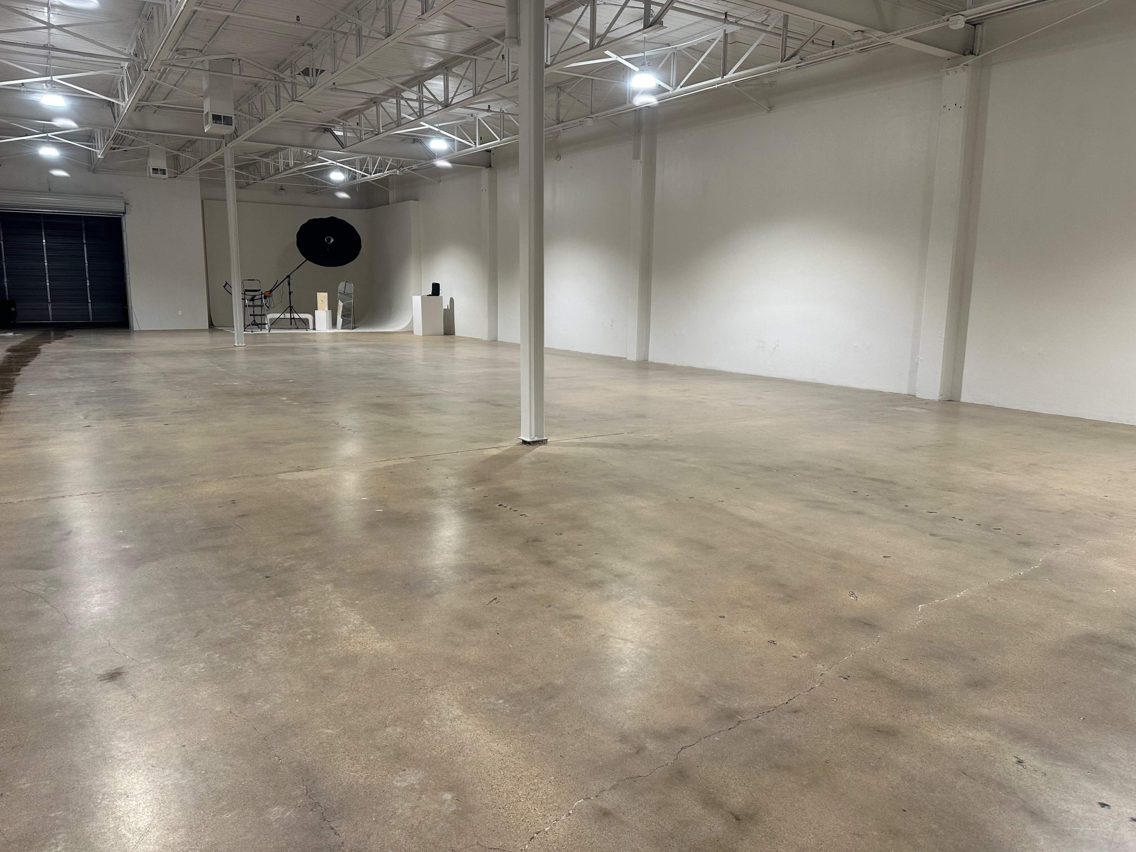 A large, empty warehouse with concrete flooring and white walls, featuring a loading bay door at the far end.