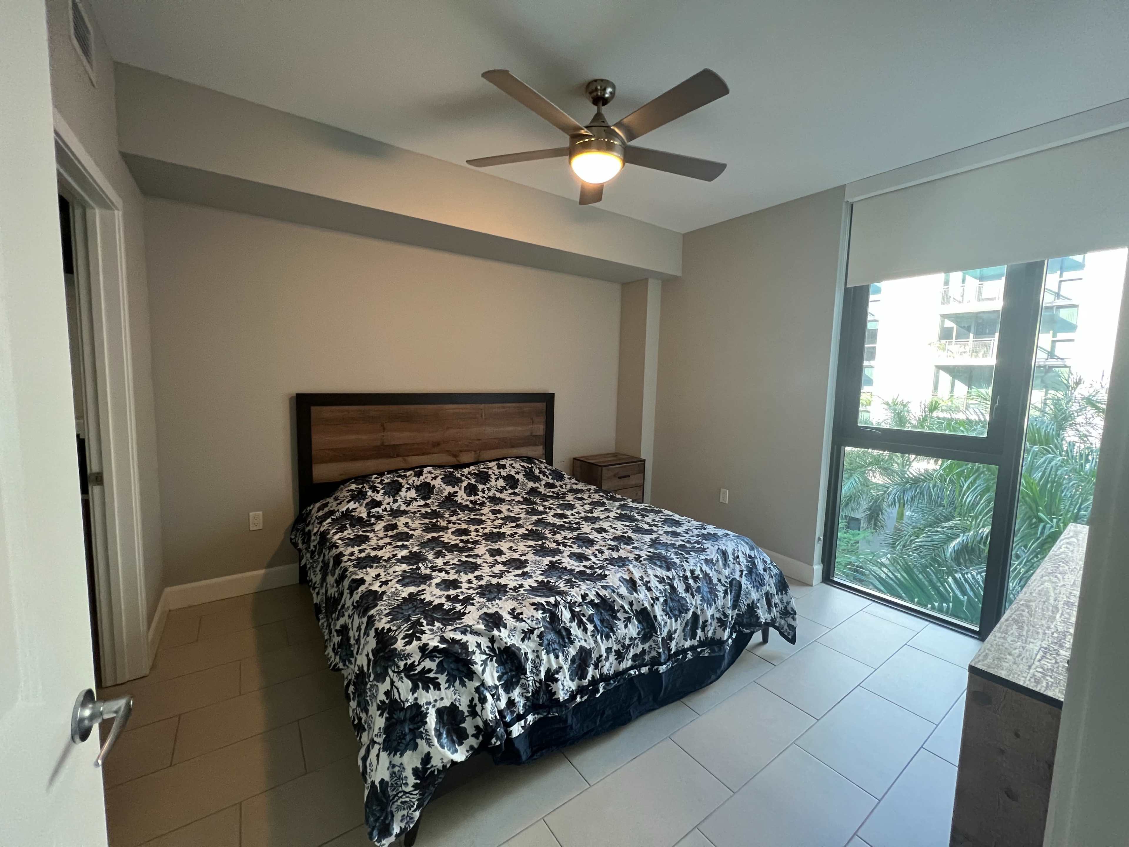 A bedroom with a large bed covered in a floral-patterned comforter, a nightstand, and a window facing greenery outside.