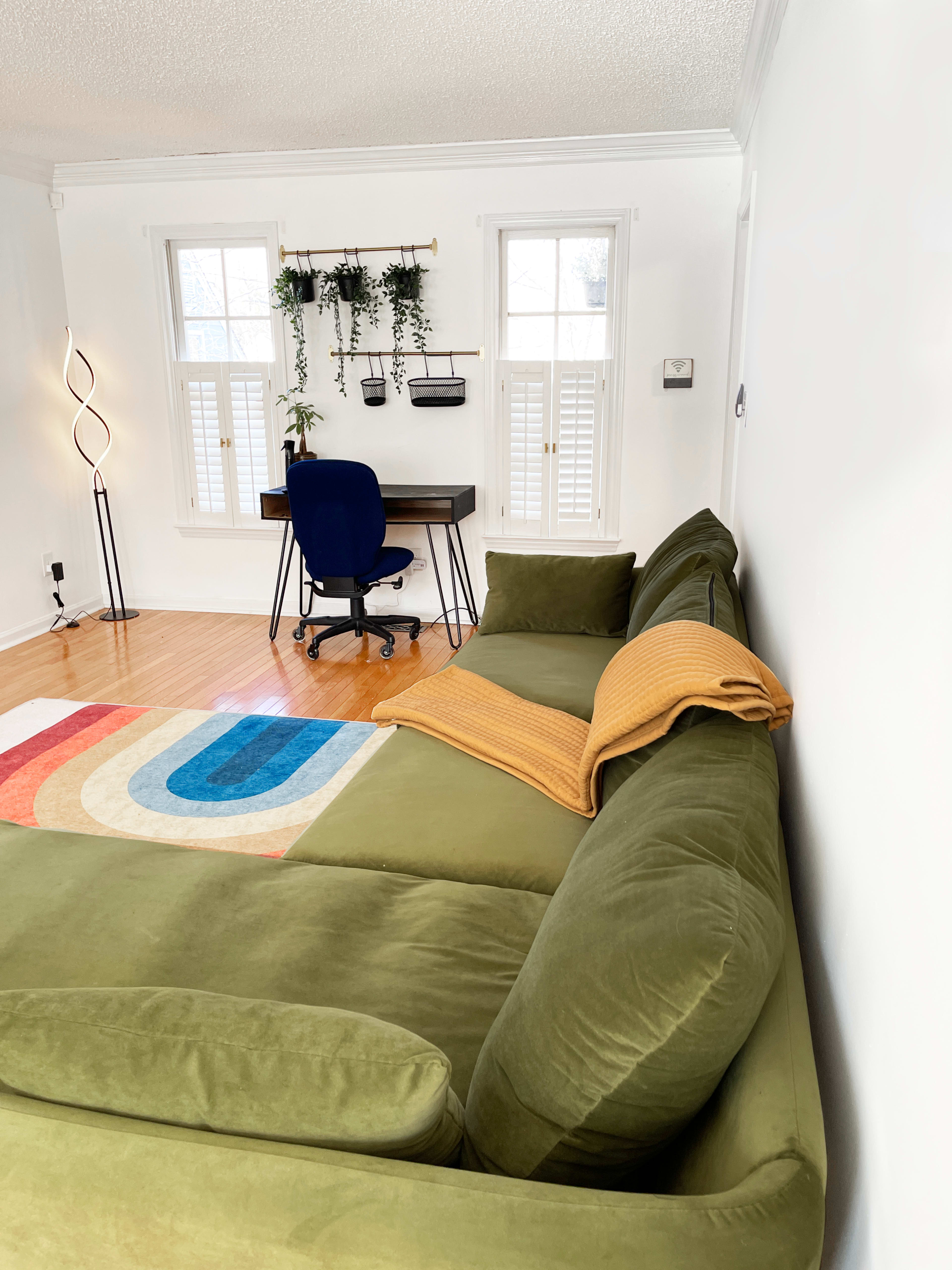 A green sectional sofa is positioned in a bright room with a wooden floor, a workspace featuring a chair and desk, and windows adorned with plants.