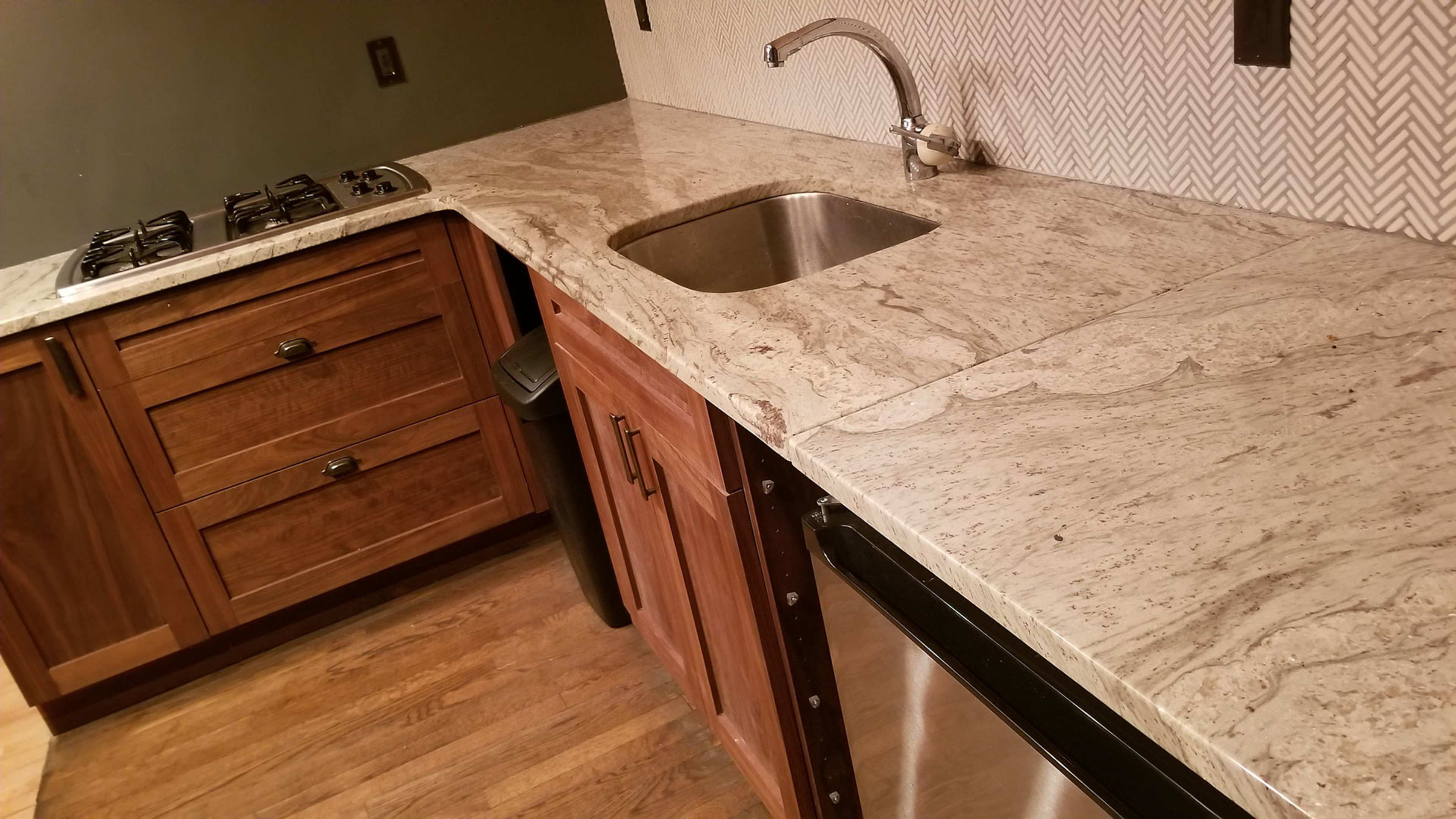 The image shows a kitchen with wooden cabinets, a stainless steel sink, and a countertop made of patterned light-colored stone.
