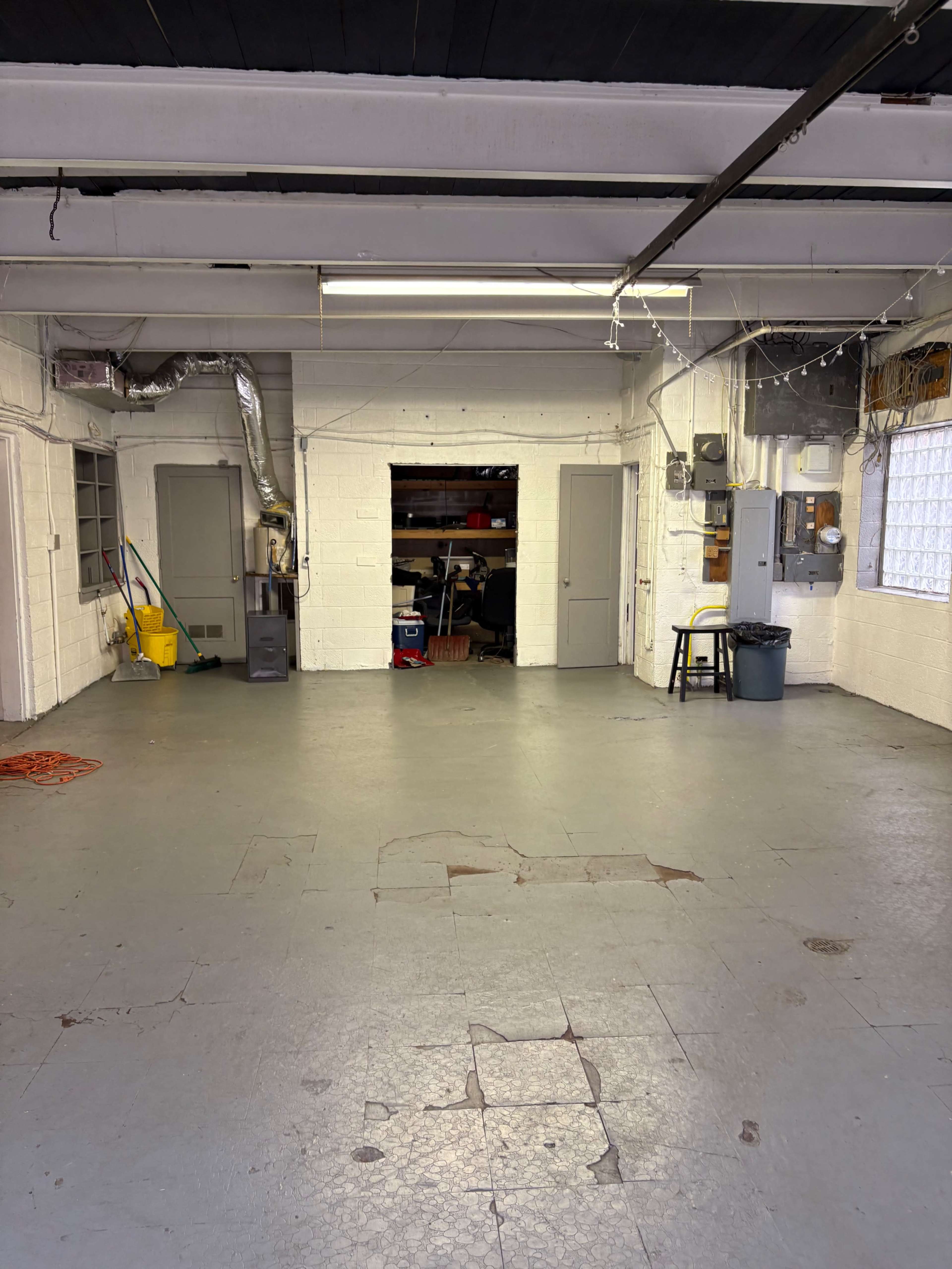 The image shows a spacious, empty garage or workshop with concrete floors, a few doors, and various utility items visible in the corners.