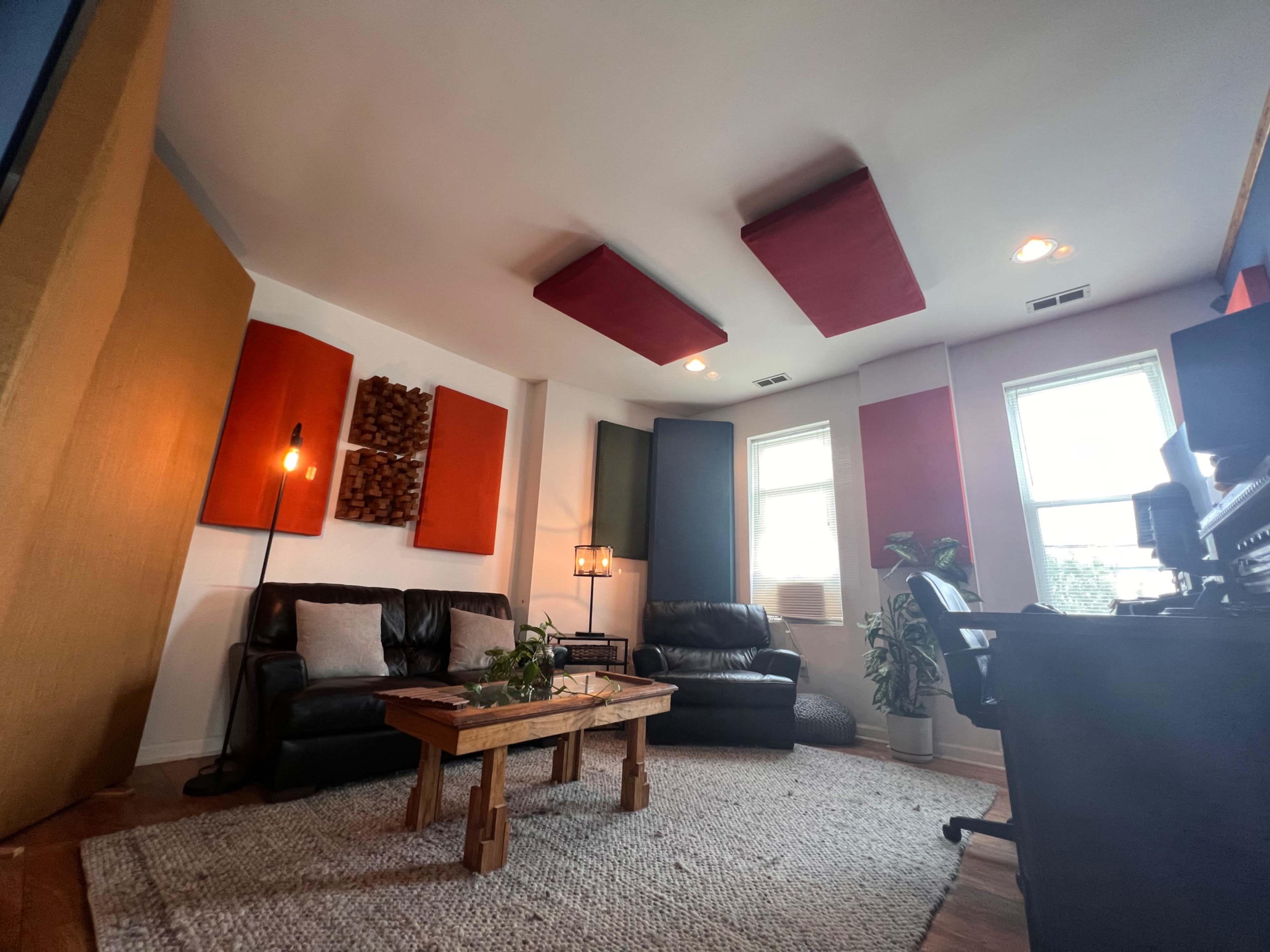 The room features two black leather sofas, a wooden coffee table, acoustic panels on the walls, and a desk with equipment in a well-lit space.