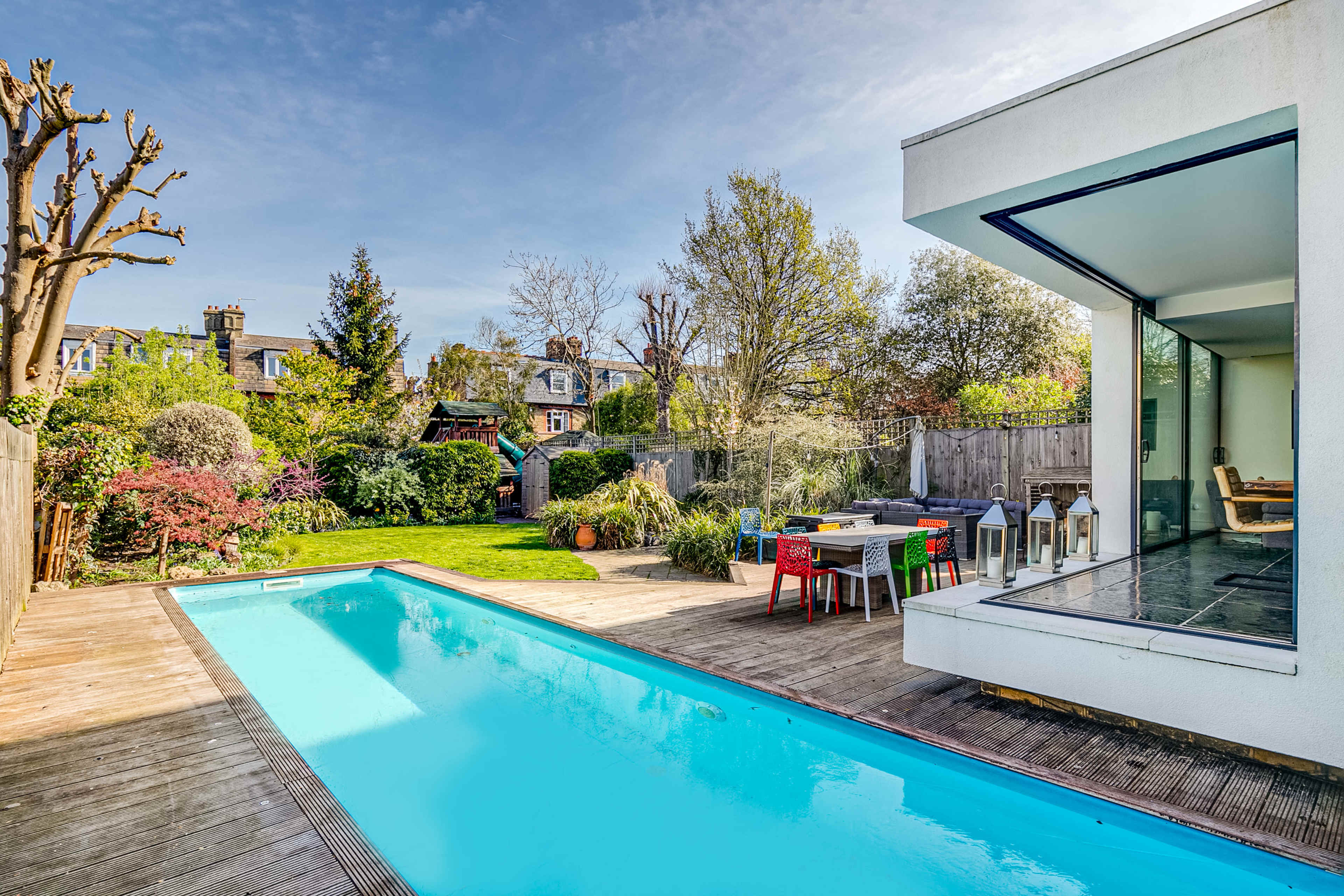 A modern home features a rectangular swimming pool surrounded by a wooden deck and a lush garden with diverse greenery and seating areas.