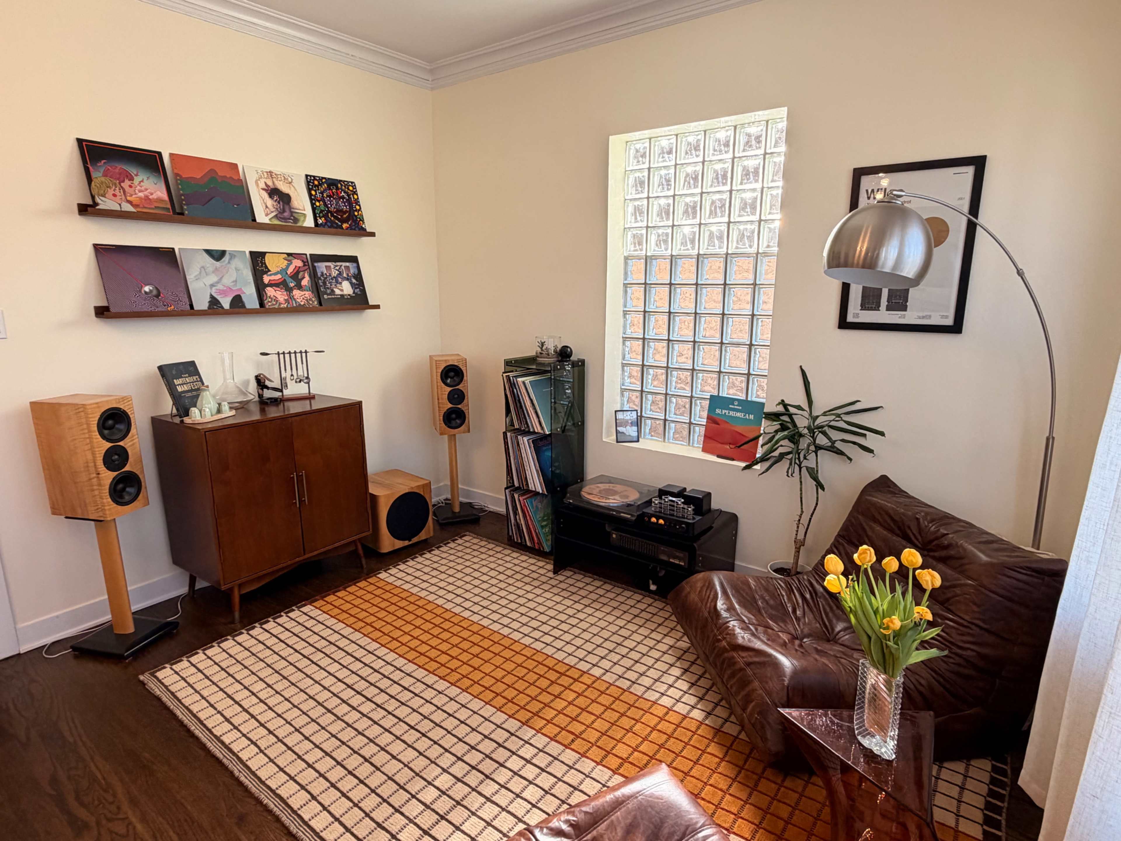 The image shows a cozy corner of a living room featuring a vinyl record shelf, a brown leather couch, a plant, and a turntable setup beside wooden speakers.