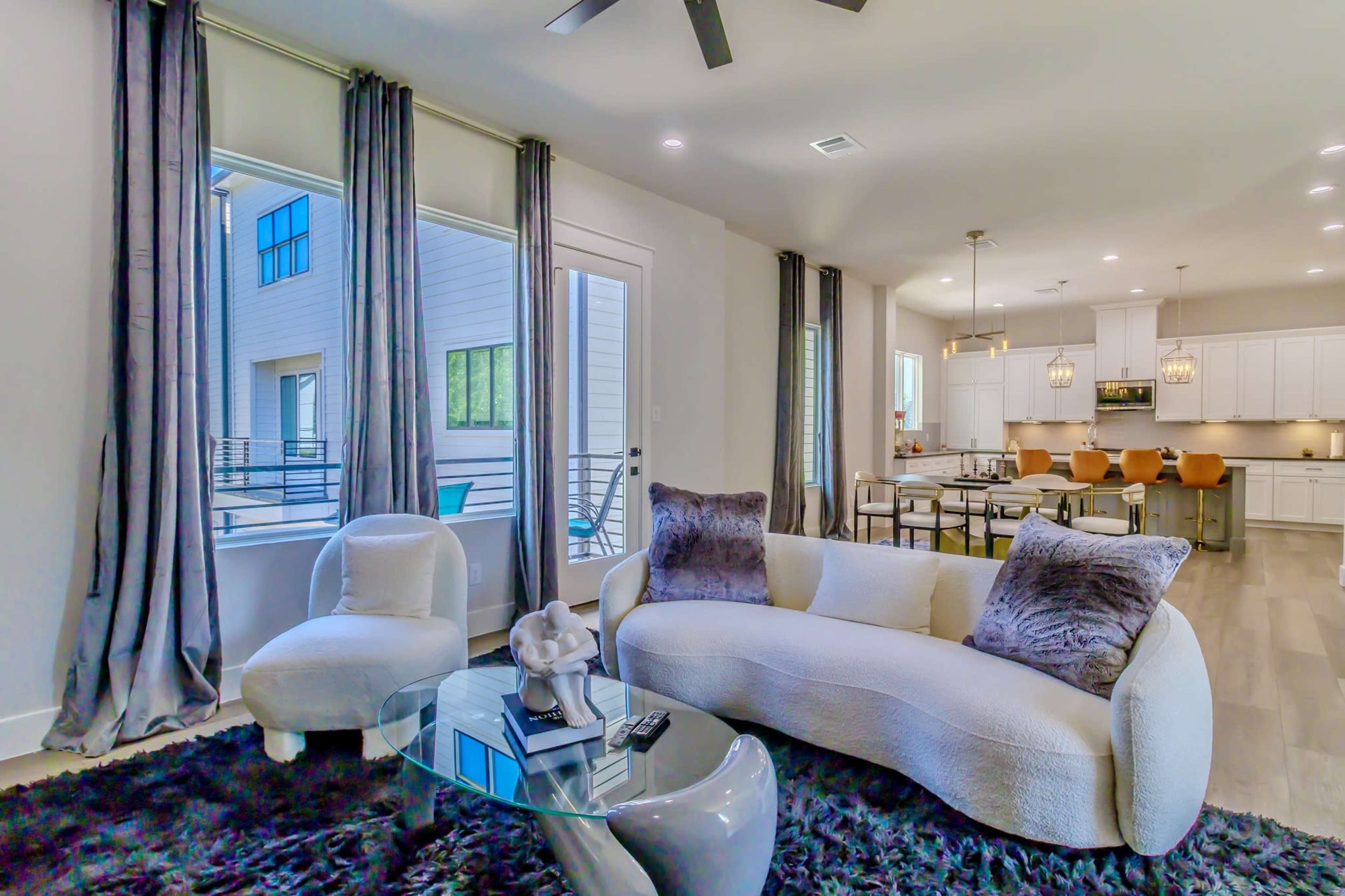 The image shows a modern living room with a white curved sofa, a glass coffee table, and large windows, leading into a kitchen area with bar stools and white cabinetry.