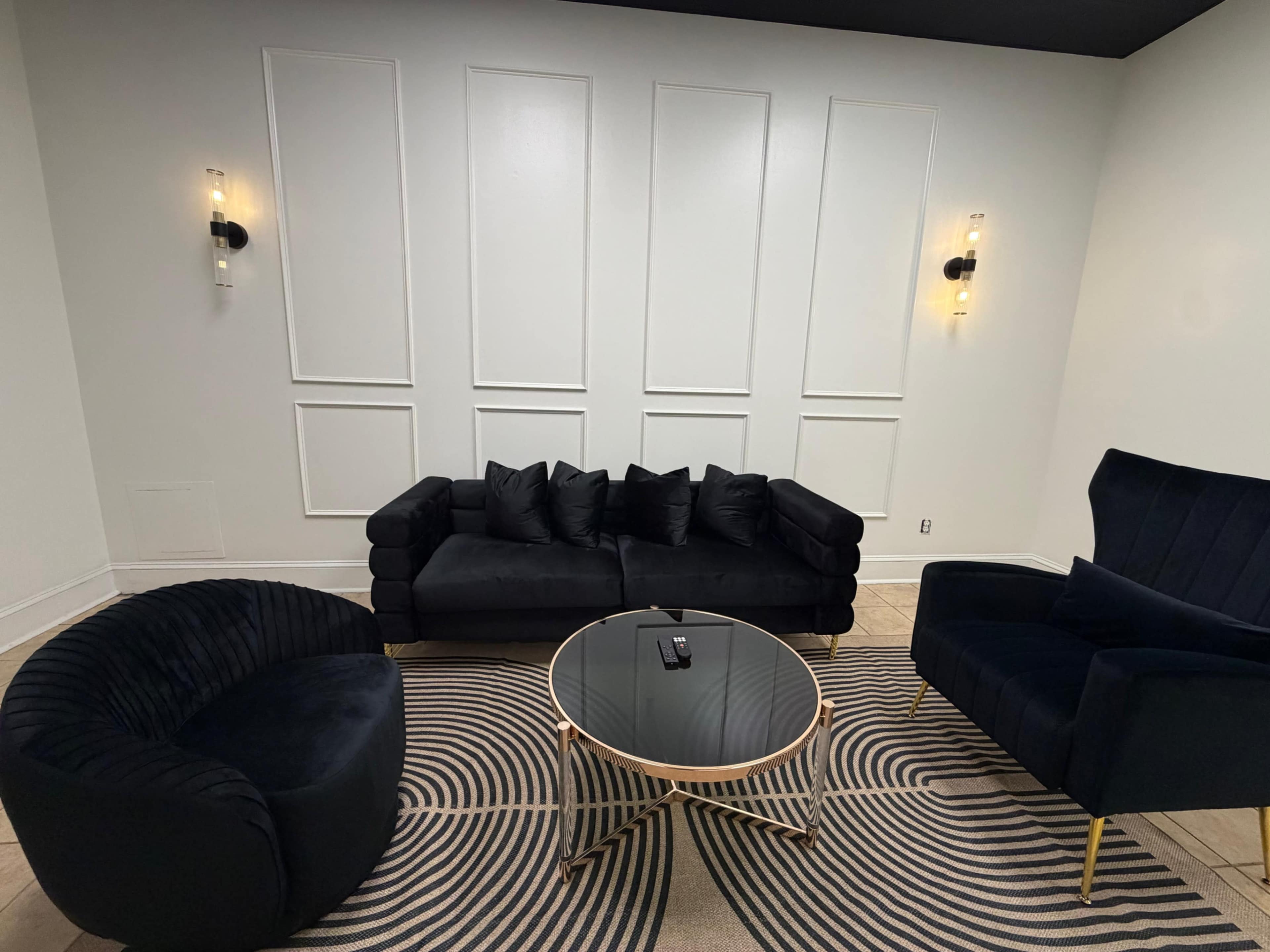 The image shows a modern lounge area featuring a black sofa, an armchair, and a round coffee table on a patterned rug.