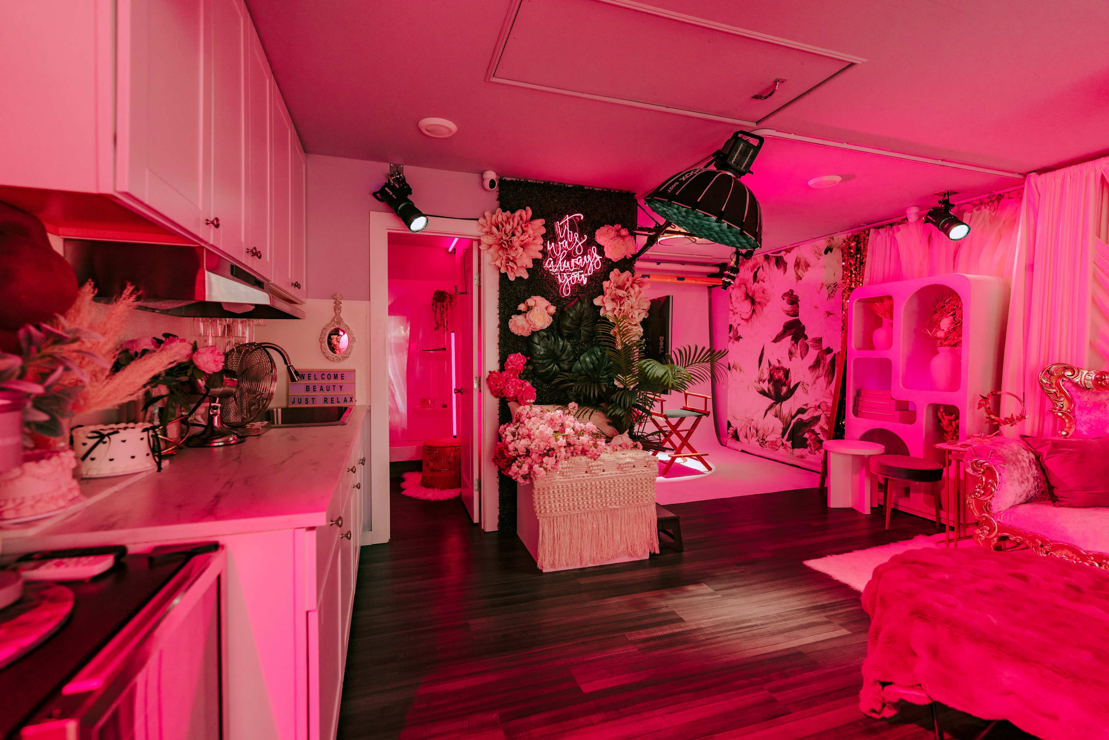 The image shows a brightly lit, modern room featuring pink lighting, floral decor, and a compact kitchen area.
