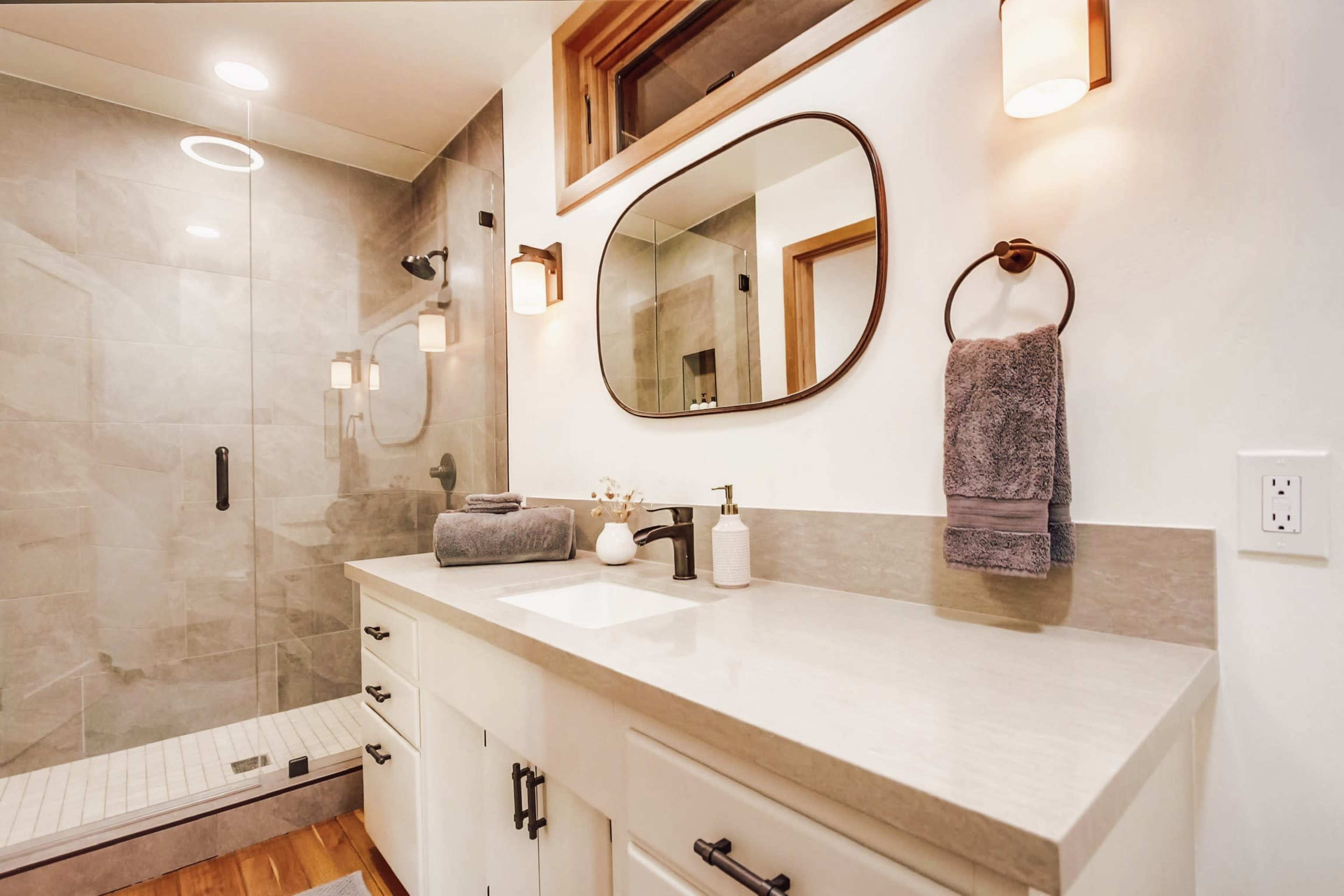 A modern bathroom features a glass shower, a round mirror above a countertop with a faucet, and neatly arranged towels and toiletries.