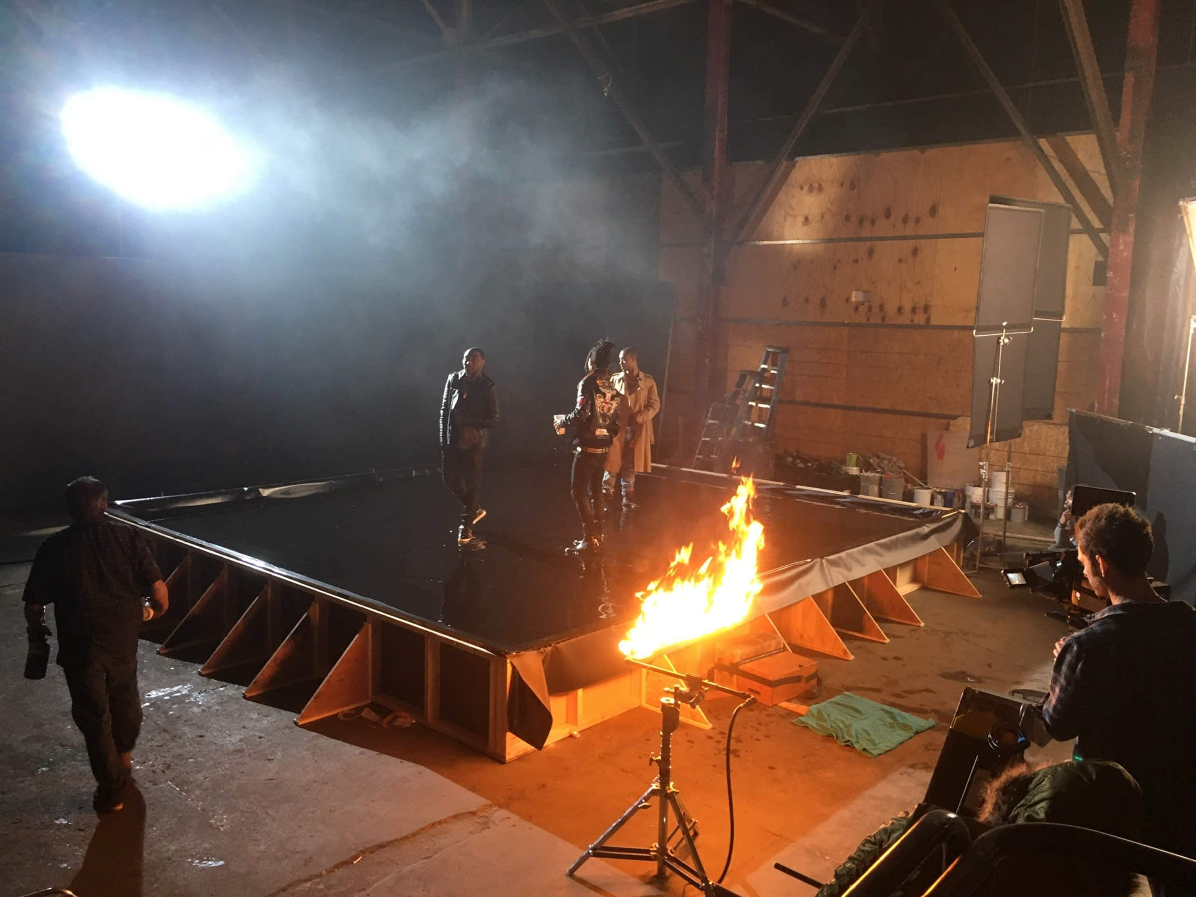 A film set features a raised platform with three individuals walking on it, while a flame burns nearby and crew members work around the scene.