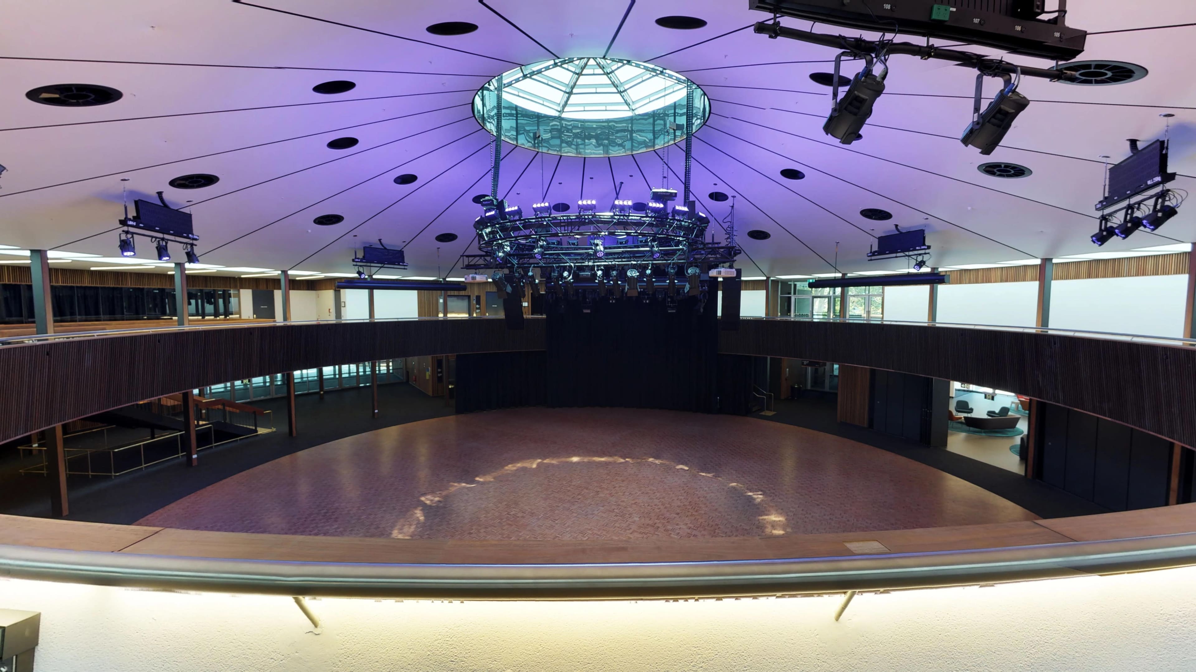 A modern auditorium with a circular stage, overhead lighting fixtures, and a glass dome ceiling, surrounded by seating areas.