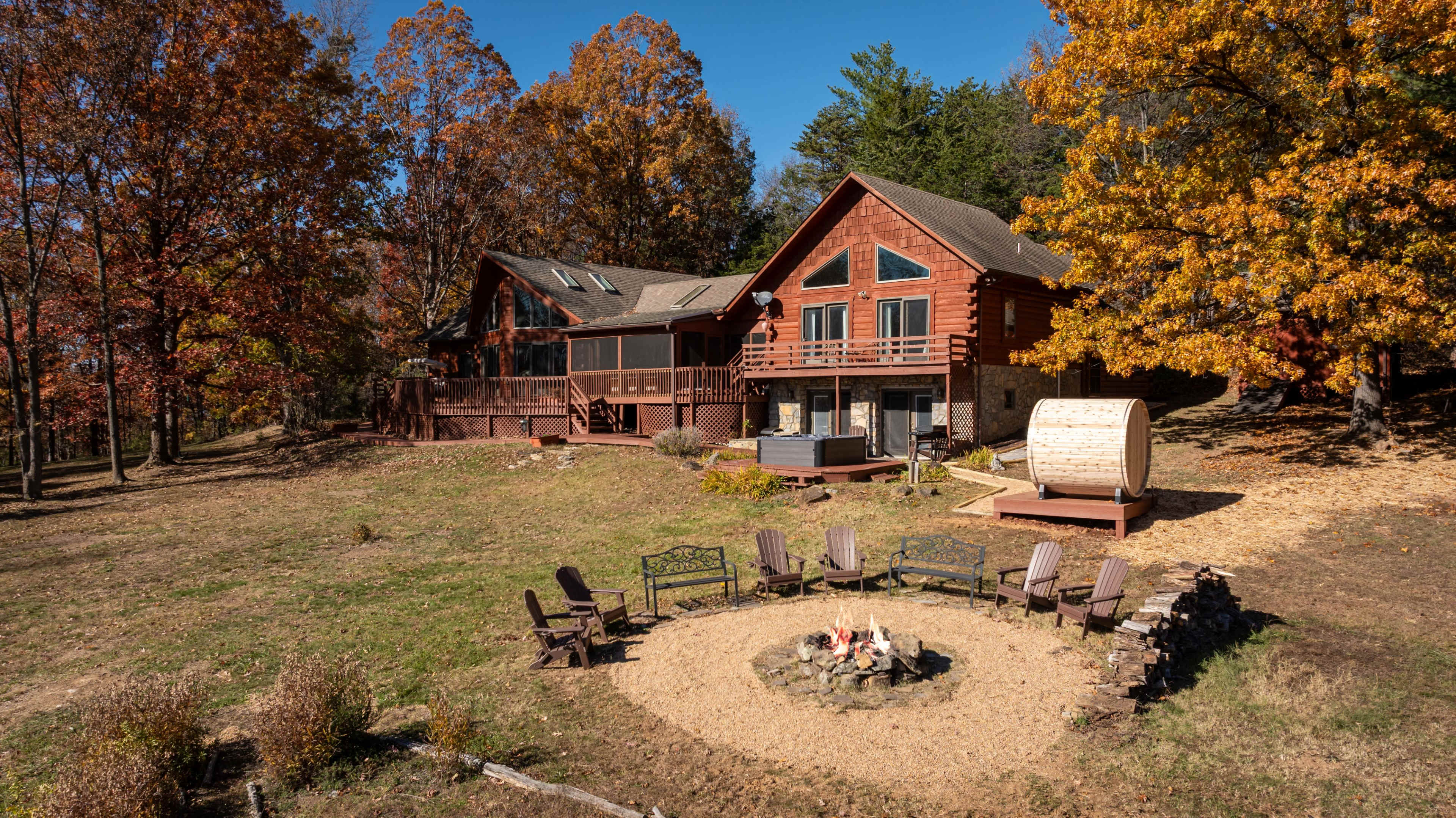 A large wooden house with a screened porch and multiple levels is surrounded by colorful autumn trees and a circular fire pit with seating.