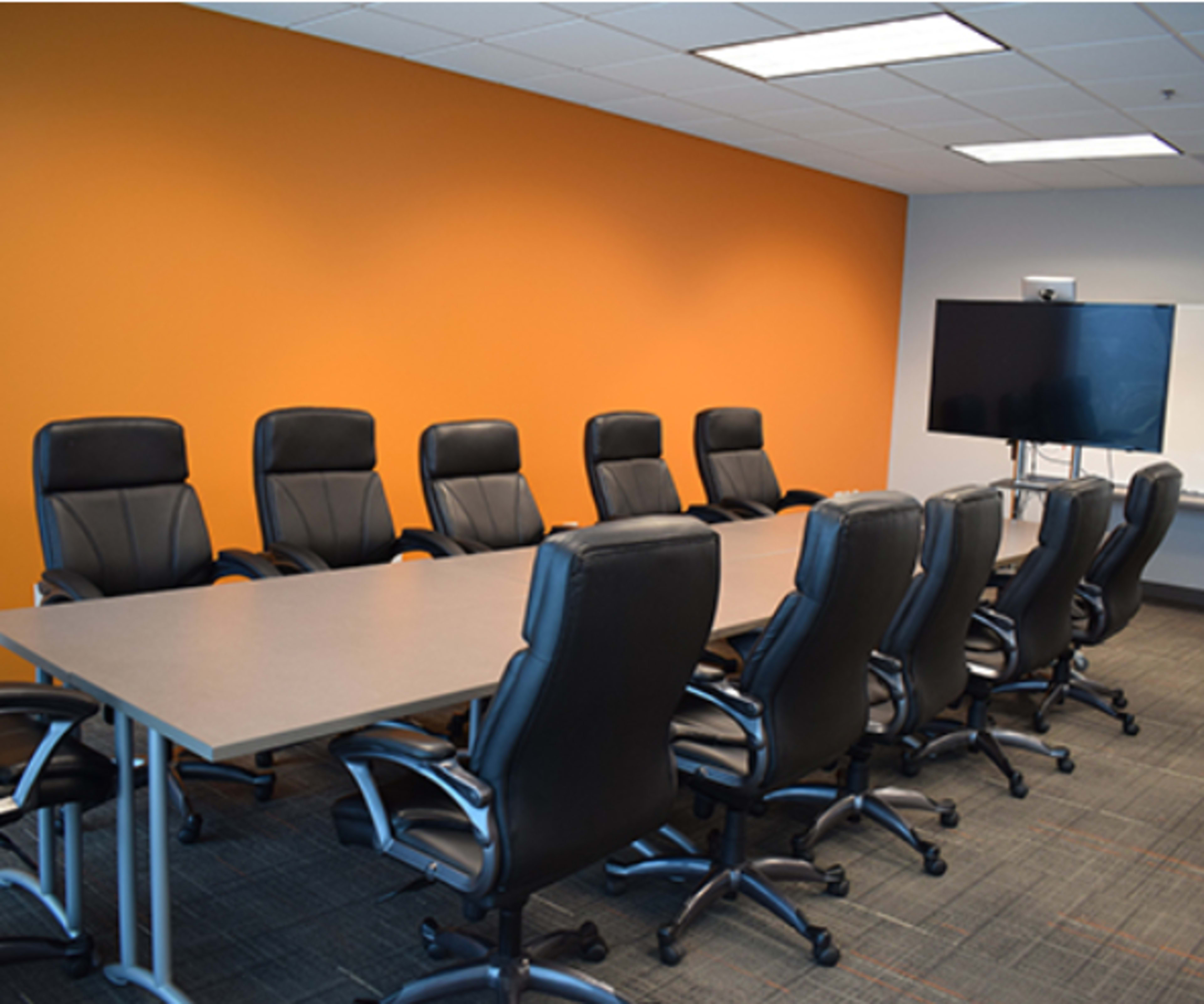 A conference room features a long table surrounded by black office chairs and an orange accent wall, with a television mounted on a stand at one end.