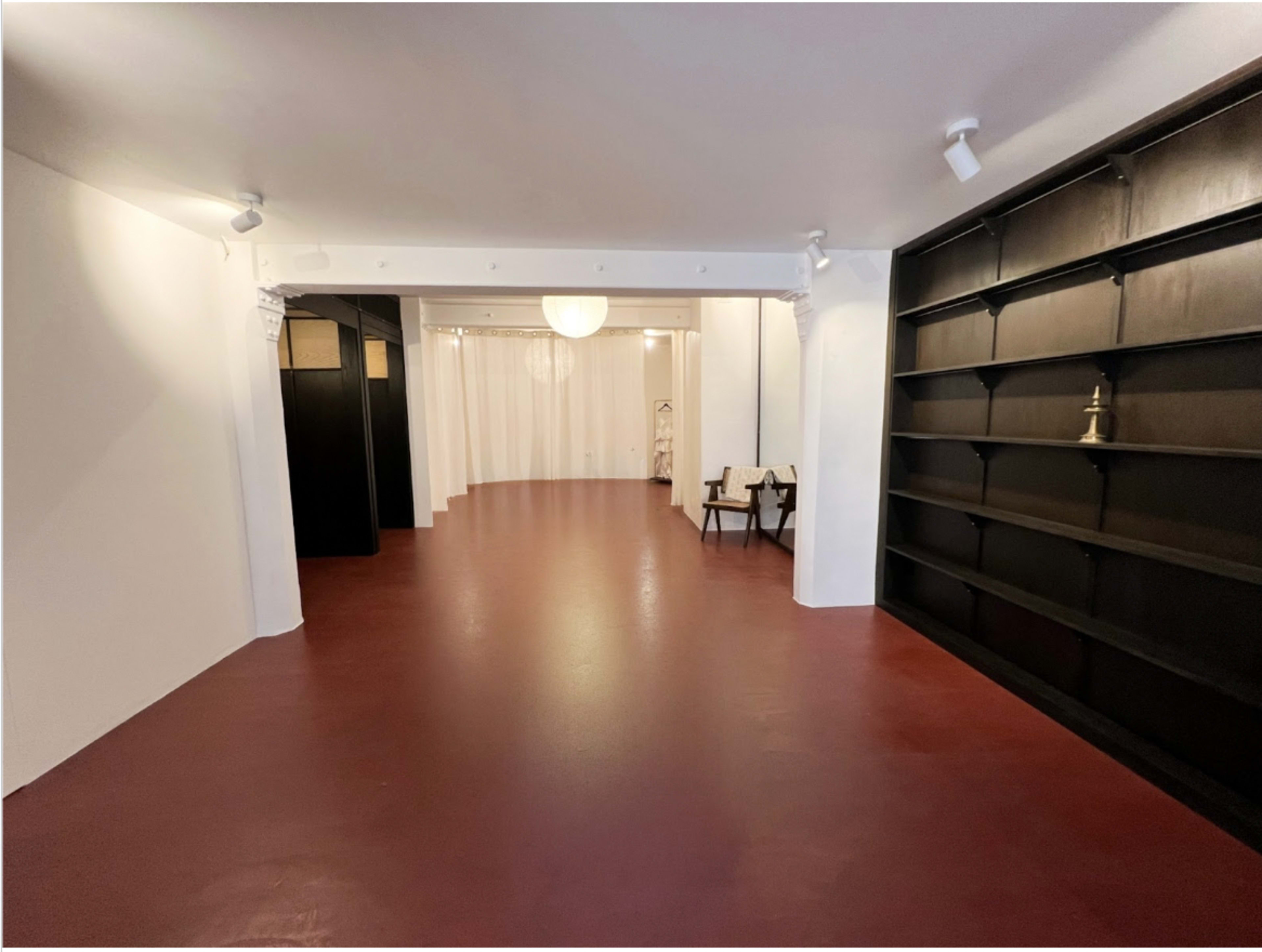 The image shows a spacious room with a red floor, white walls, a large light fixture, and bookshelves on one side.