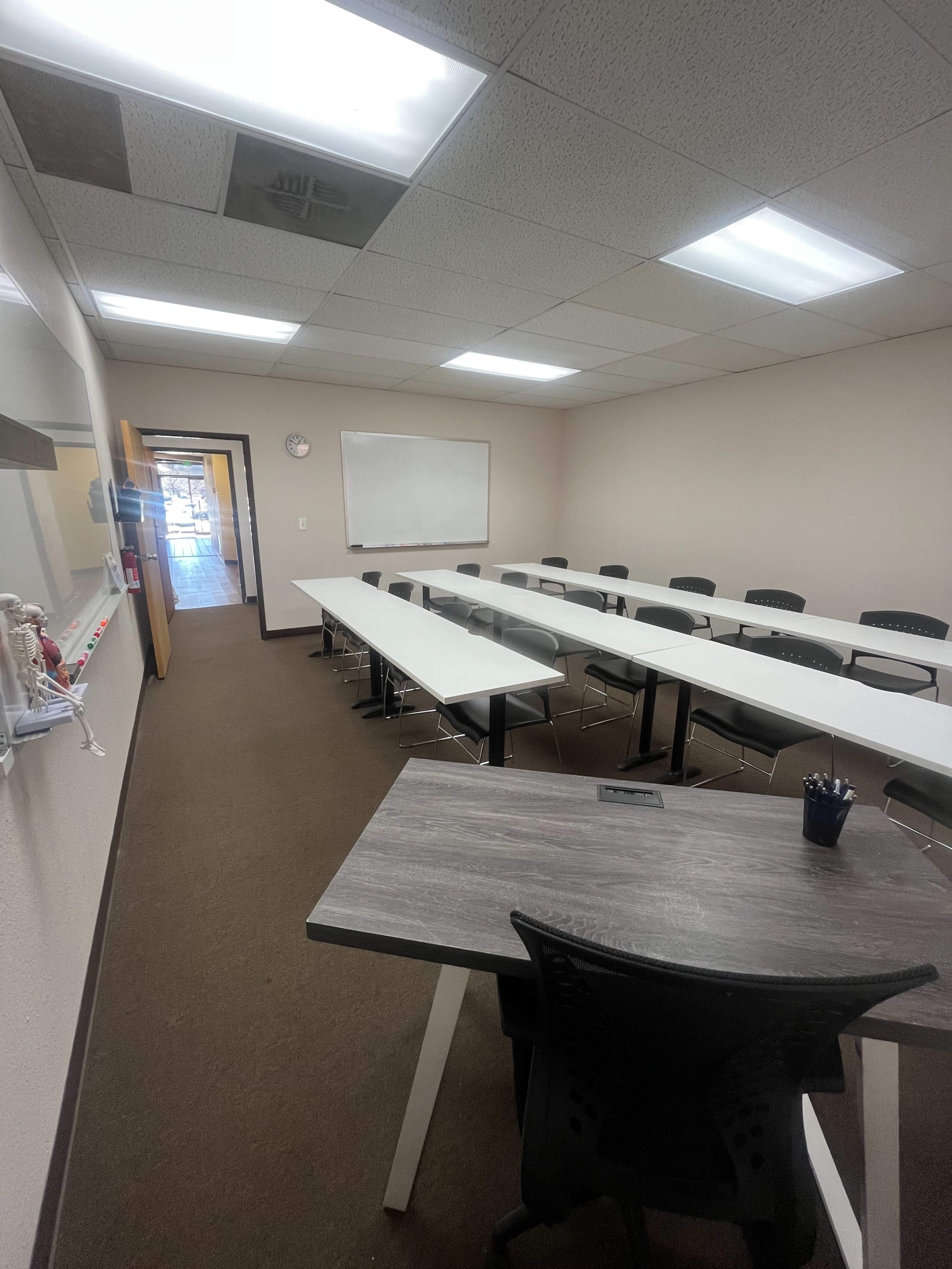 A classroom is arranged with long tables and chairs, featuring a whiteboard, clock, and an entrance at the far end.