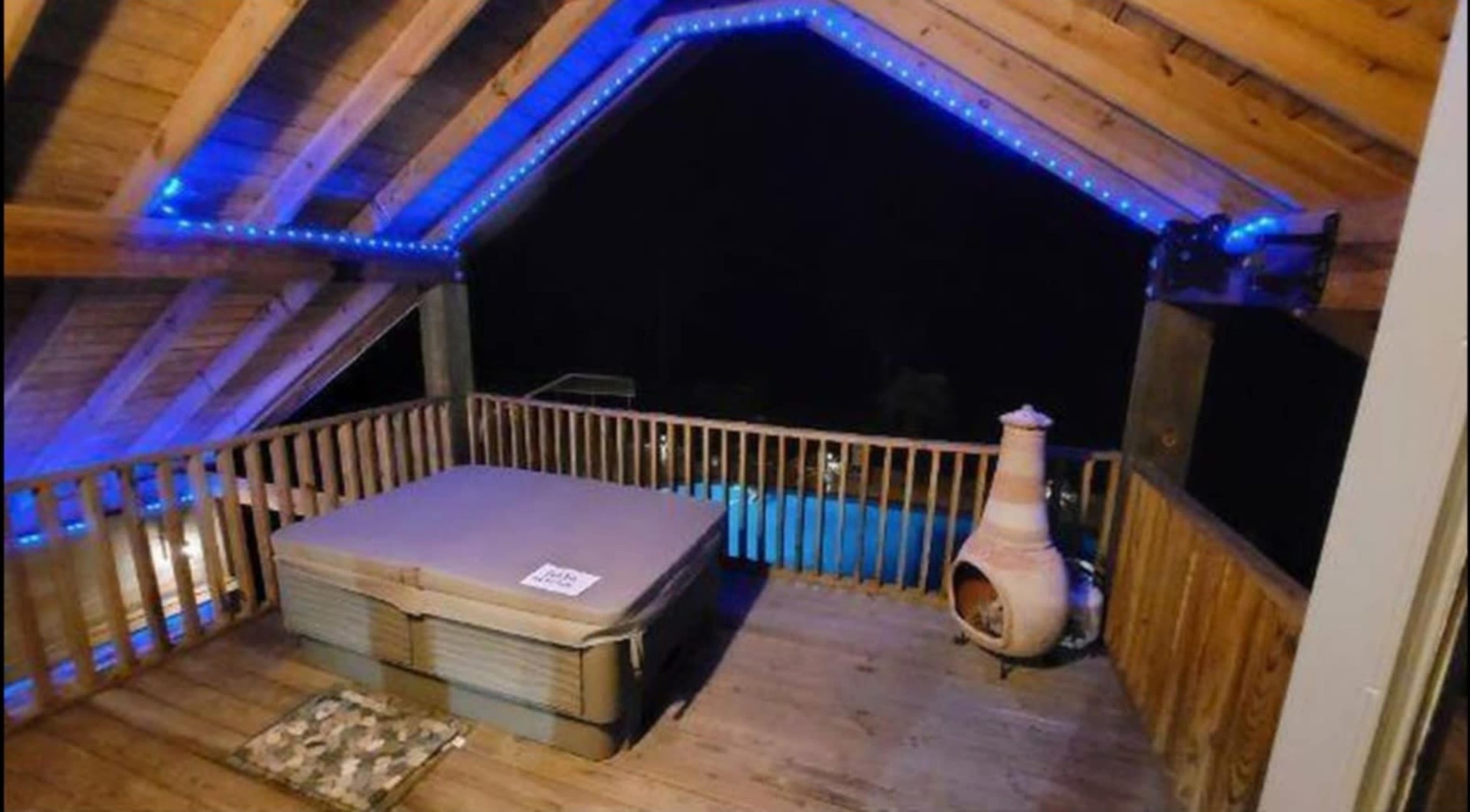 A wooden terrace with a hot tub, blue lights along the ceiling, and a pool visible in the background at night.