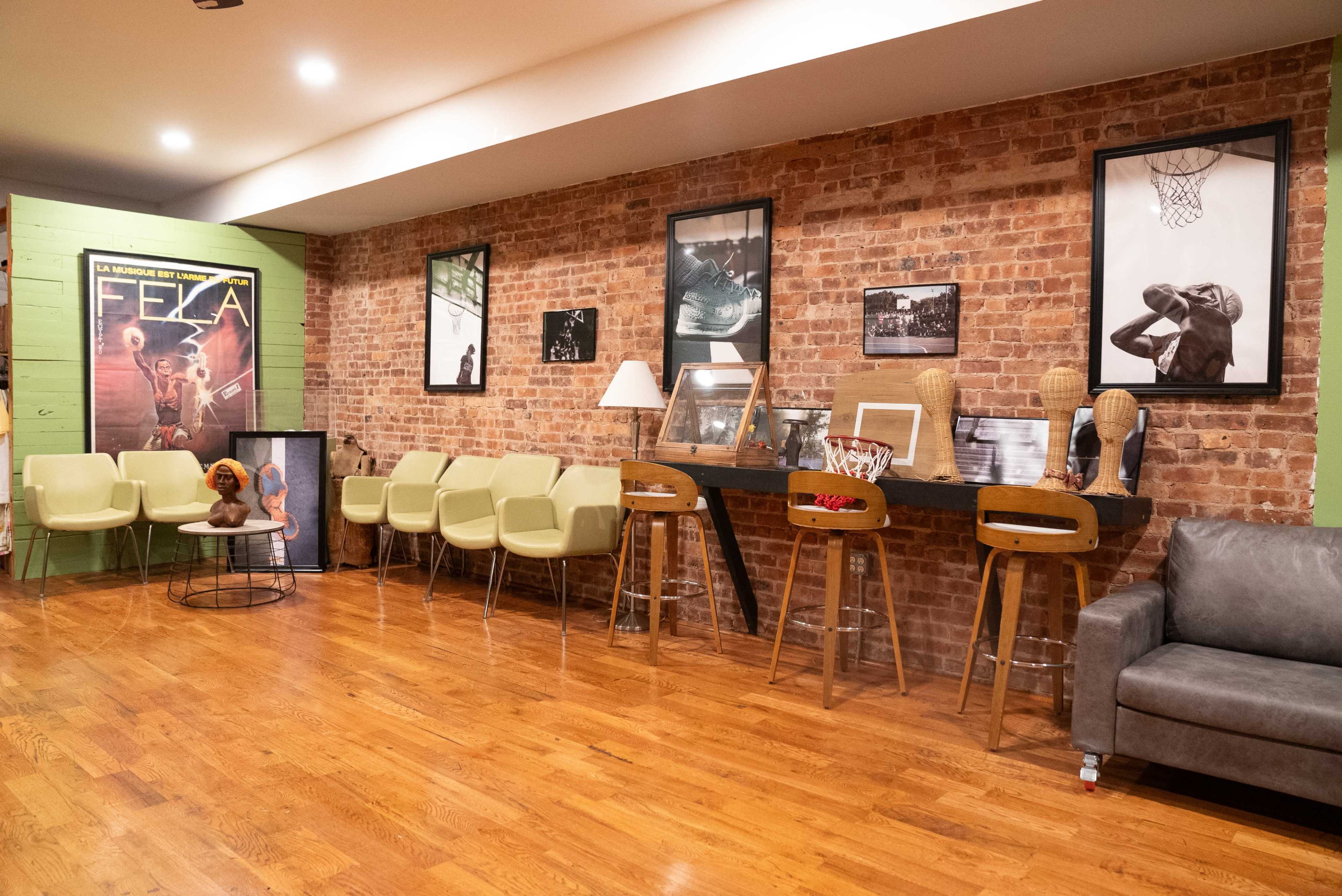 The image shows a cozy indoor space with a brick wall, featuring seating along one side, a bar-like table with stools, and various framed photographs and art pieces displayed on the walls.