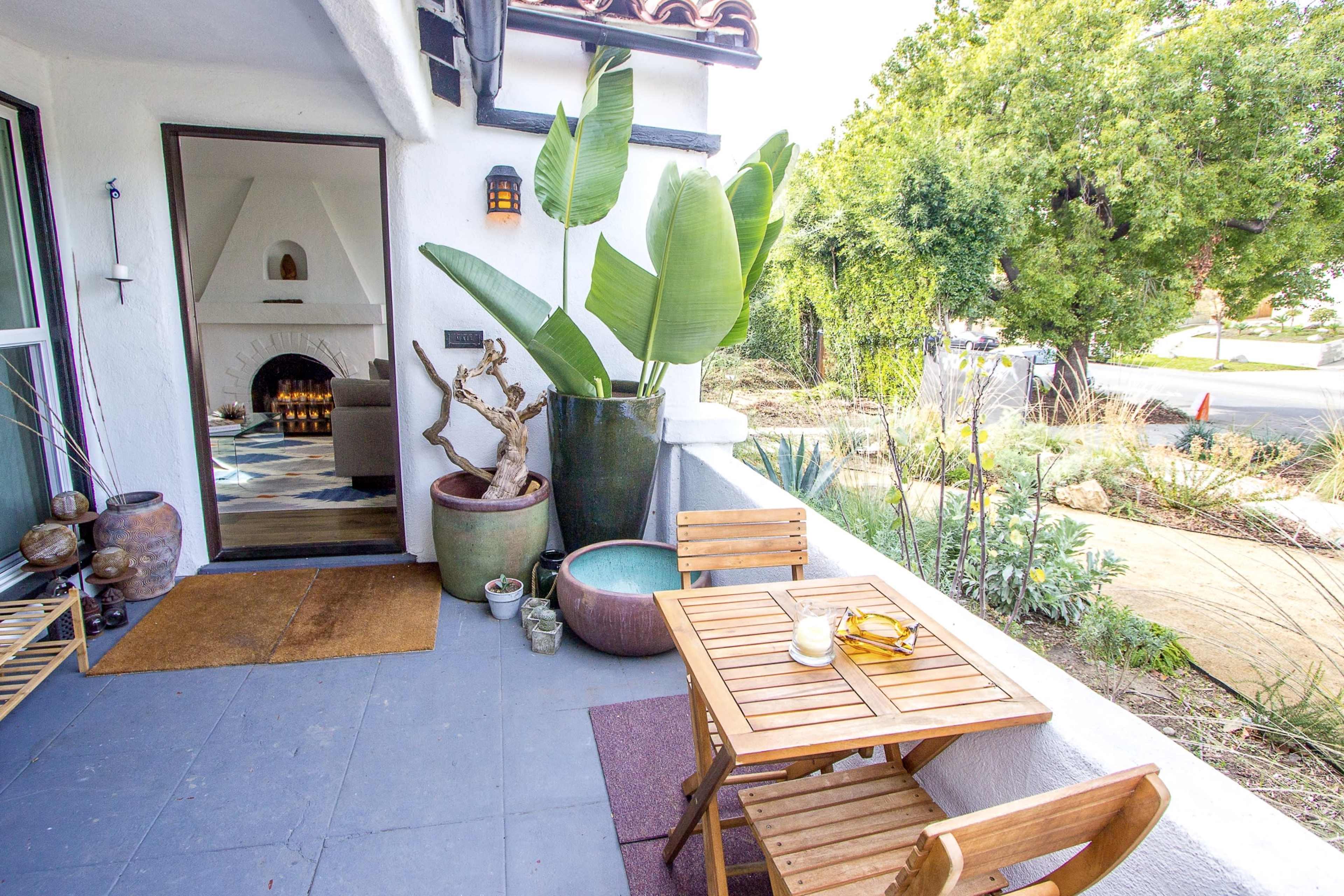 A small wooden table and chairs are set on a porch surrounded by potted plants and a view of a garden.