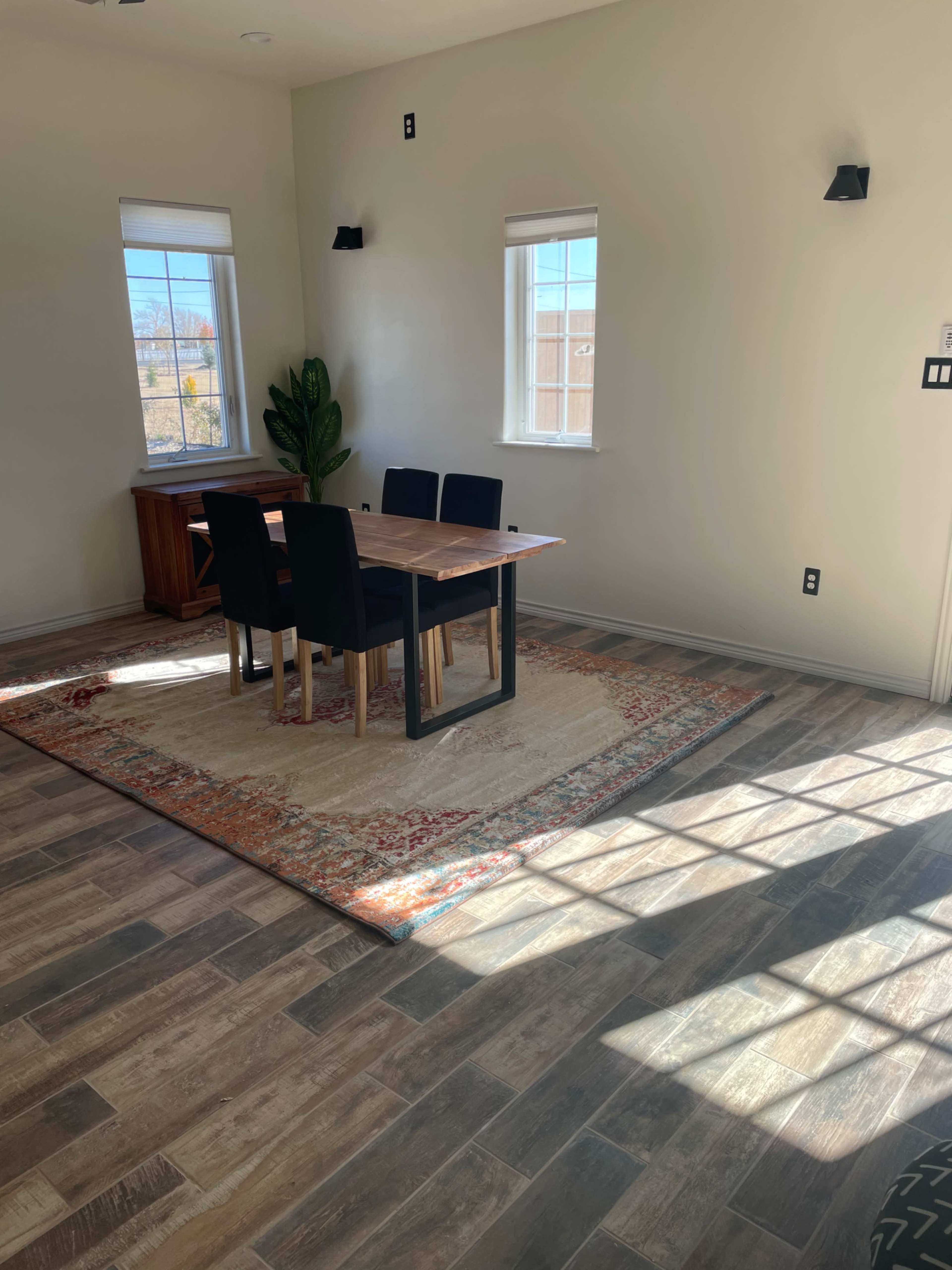 A dining area with a wooden table and four chairs set on a patterned rug, surrounded by windows letting in natural light.