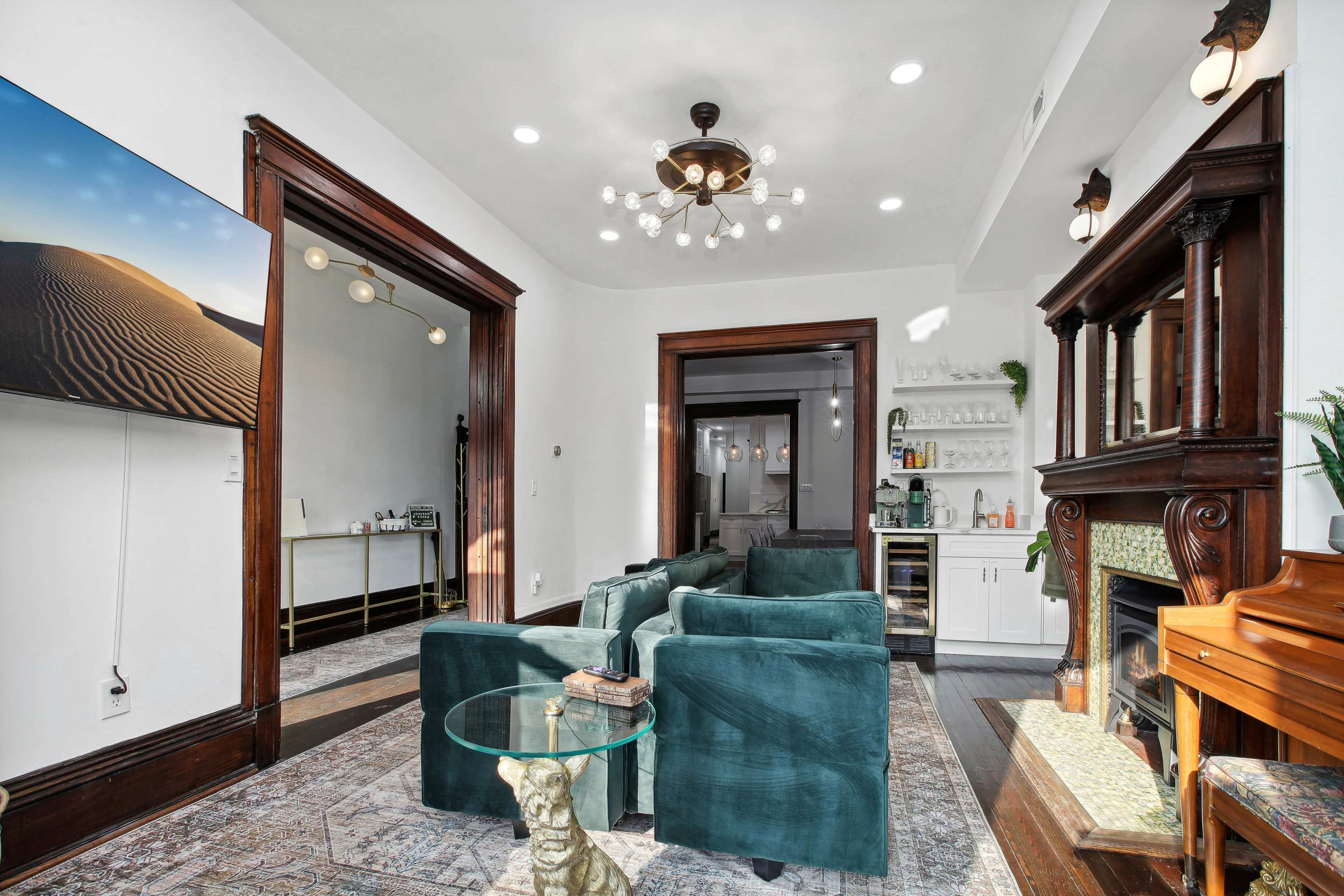 The image shows a bright living room featuring a seating area with green upholstered chairs, a glass table, and a decorative fireplace, alongside a piano and a large wall-mounted screen displaying a desert landscape.