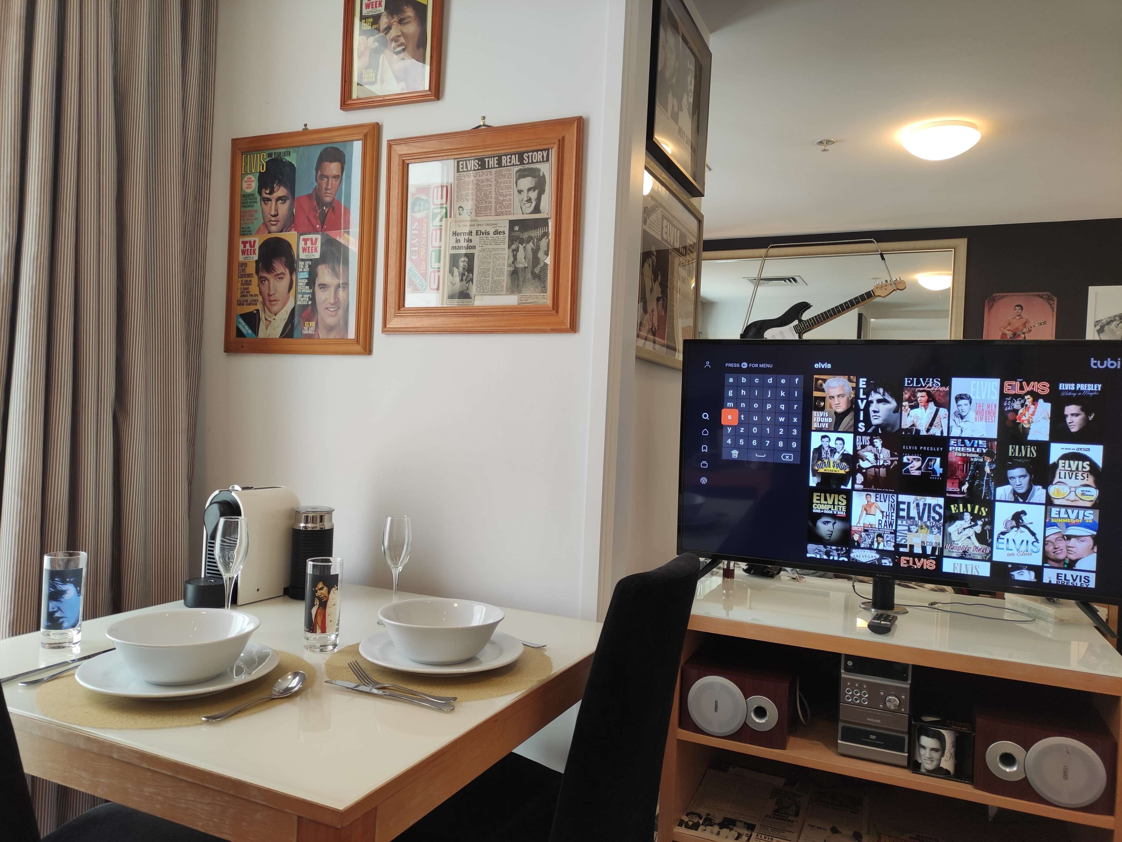 A dining area with a table set for two, featuring bowls and utensils, alongside a television displaying a menu of Elvis-related content, surrounded by framed photographs on the wall.