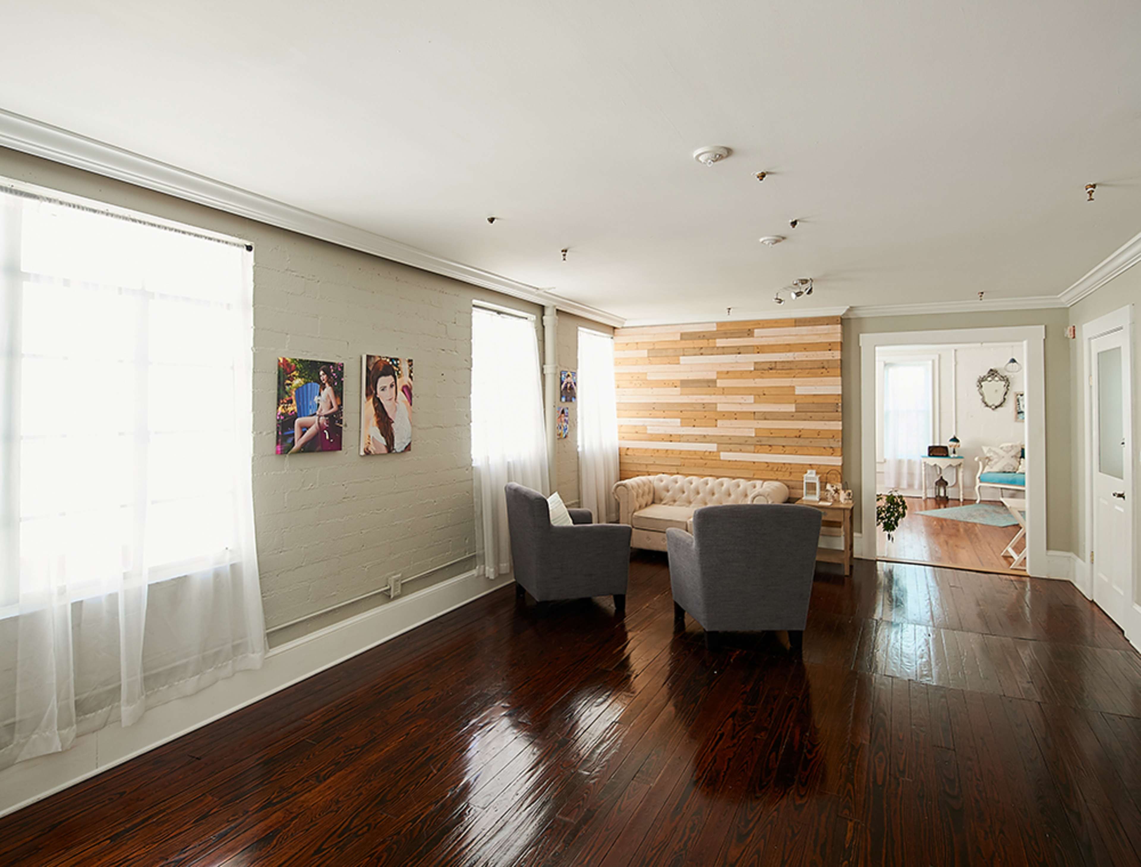 The image shows a well-lit room with a cozy seating area featuring two gray chairs and a wall made of wooden panels, accompanied by large windows with sheer curtains.