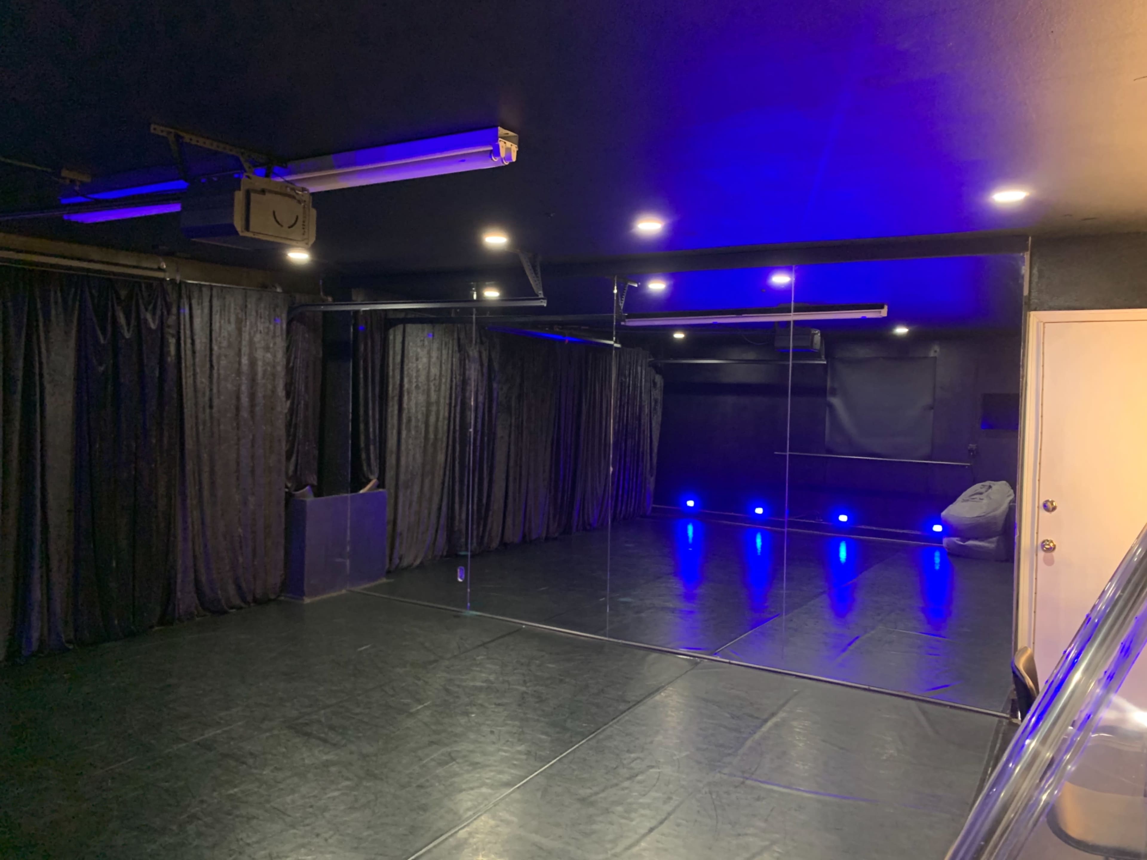 A spacious dance studio with a black floor, mirrors on one wall, black curtains, and blue LED lighting.