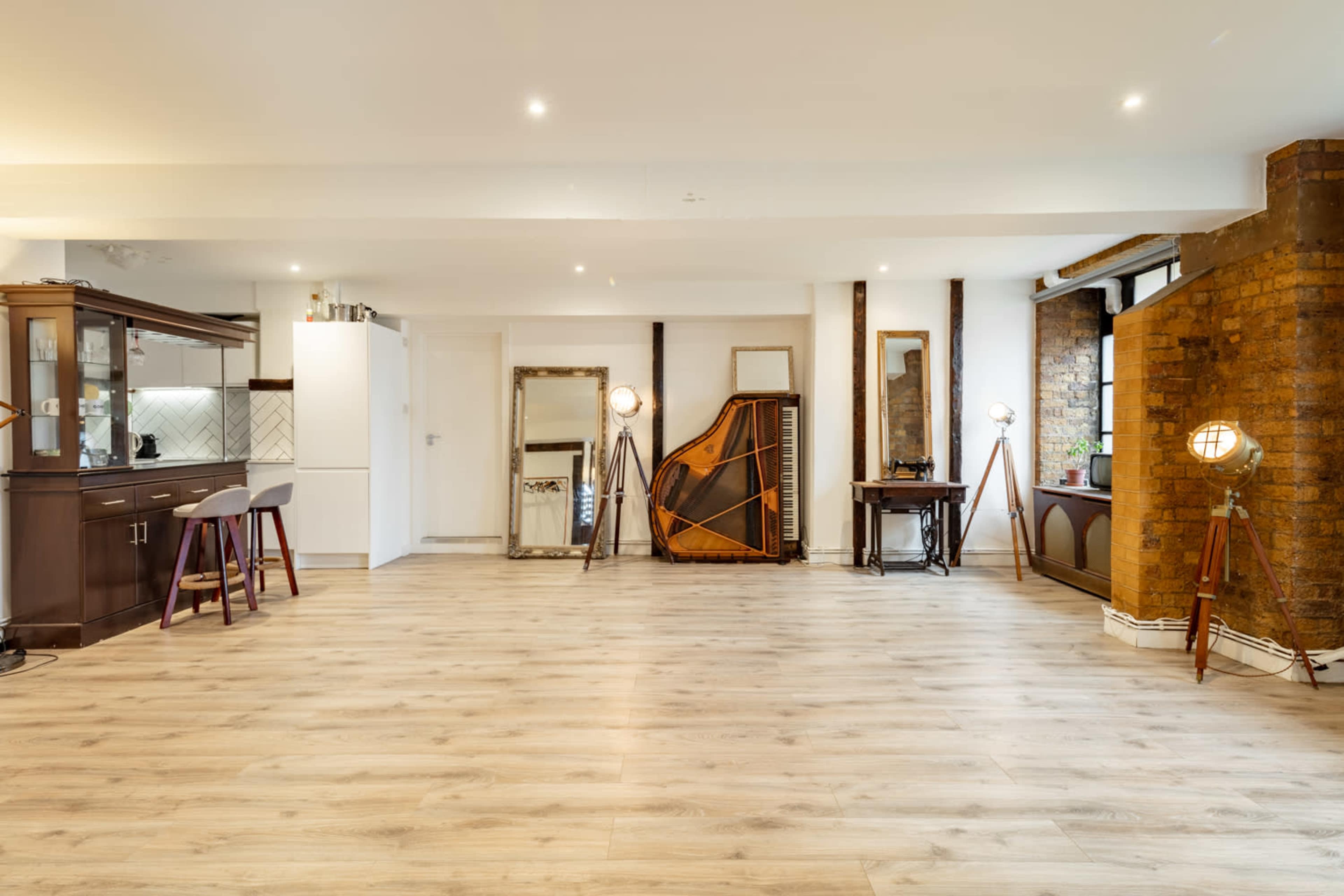 A spacious, well-lit room featuring wooden flooring, a kitchen area with cabinets, a large mirror, a vintage-style harp, and decorative floor lamps.