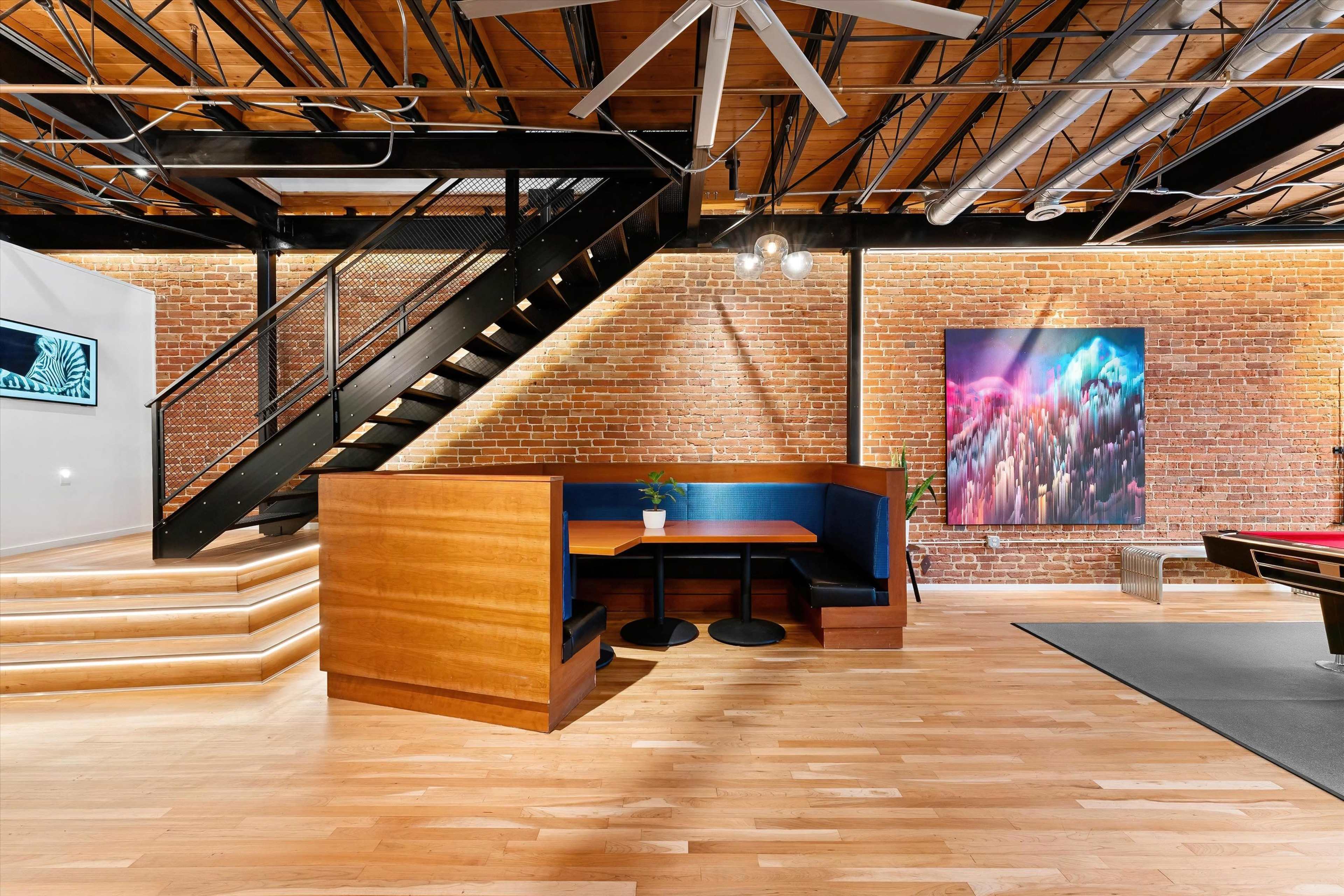 A modern interior space featuring a wooden staircase, a seating area with a booth and tables, exposed brick walls, and a colorful artwork on one wall.