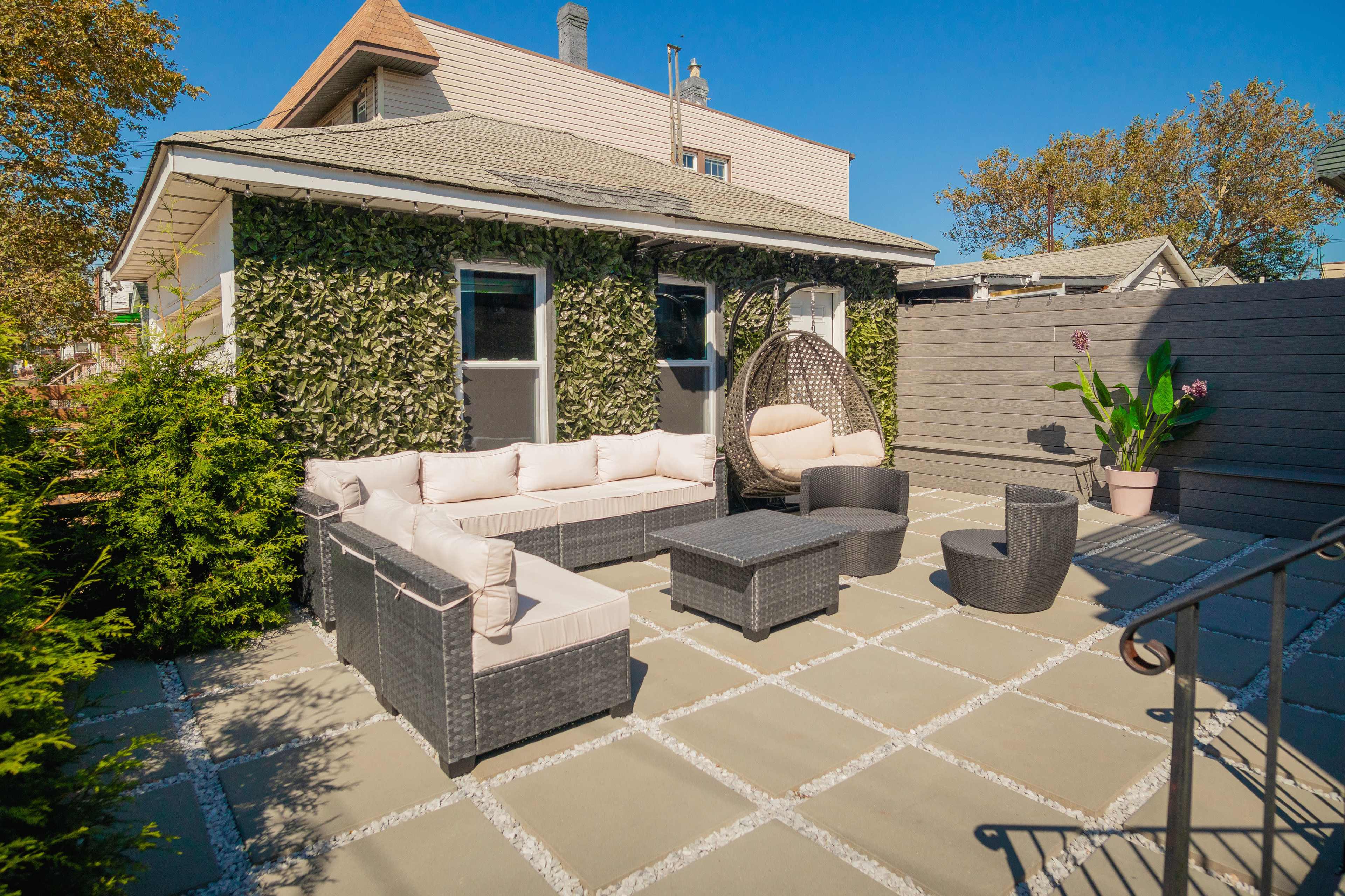 A patio area features a sectional sofa, a hanging chair, and a coffee table surrounded by potted plants and a wall covered in greenery.