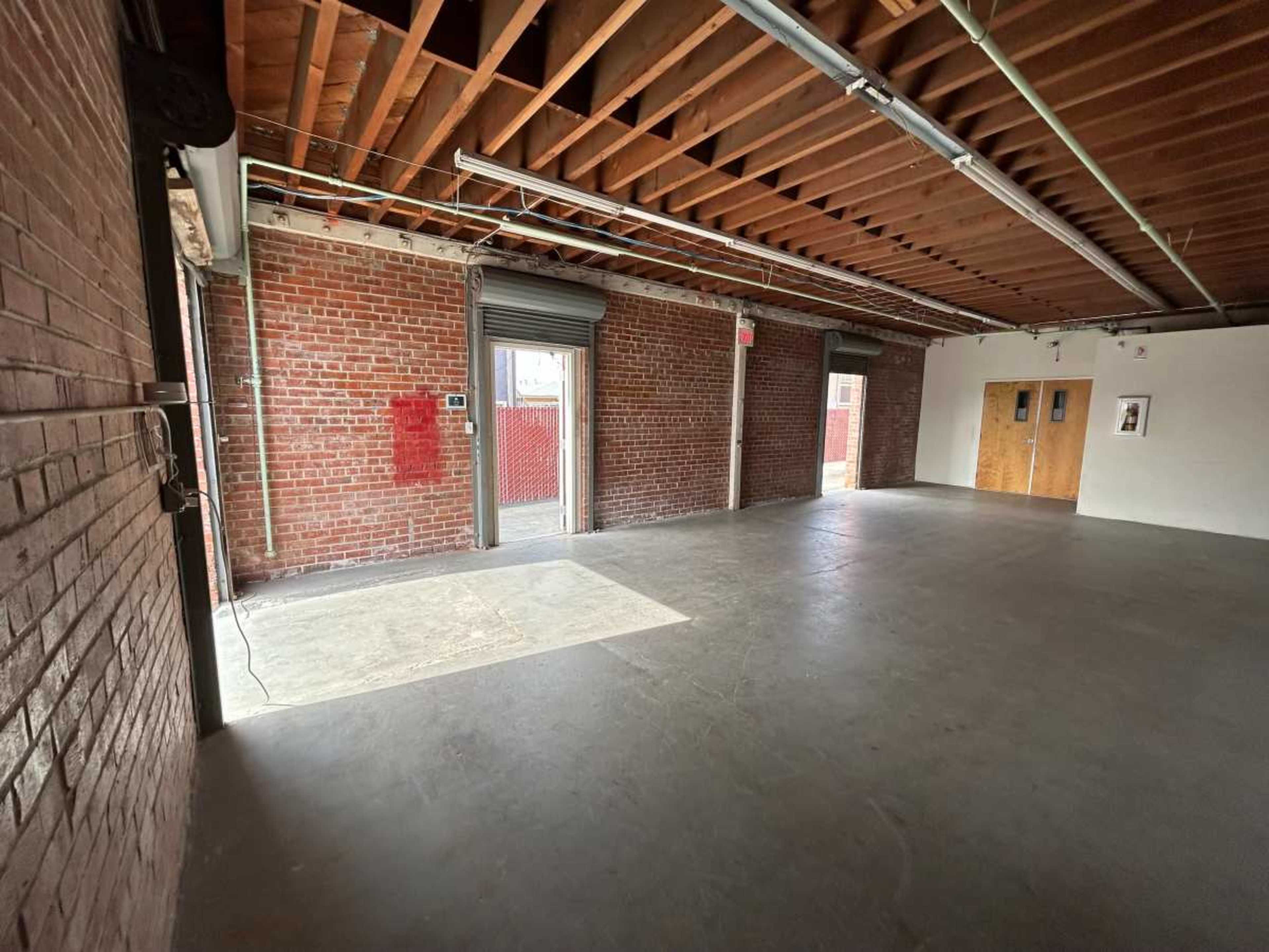 An empty industrial space with exposed brick walls, a concrete floor, and large doors leading to an outdoor area.
