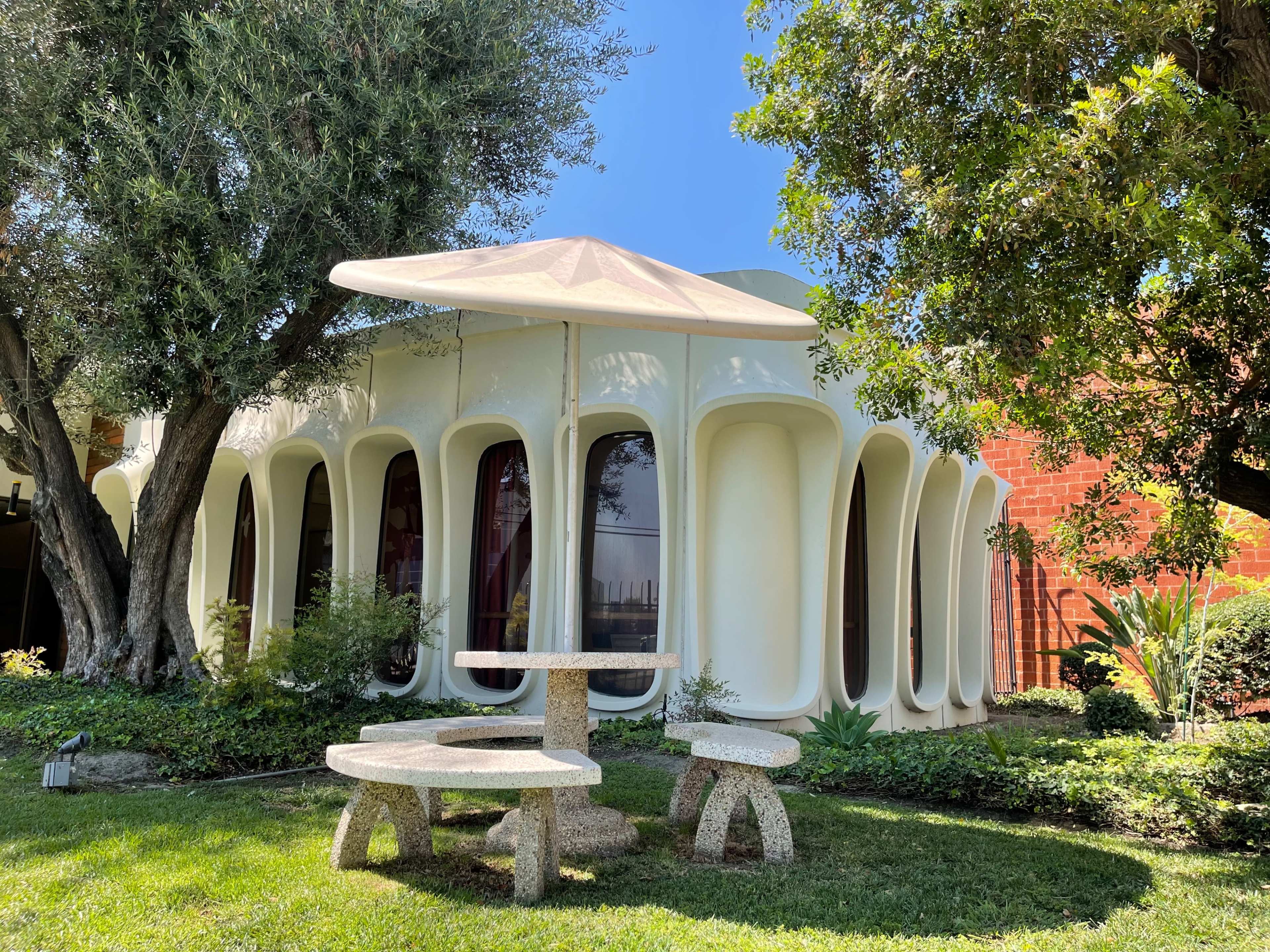 The image shows a modern white building with unique arching columns, surrounded by lush greenery and a circular stone table under a canopy.