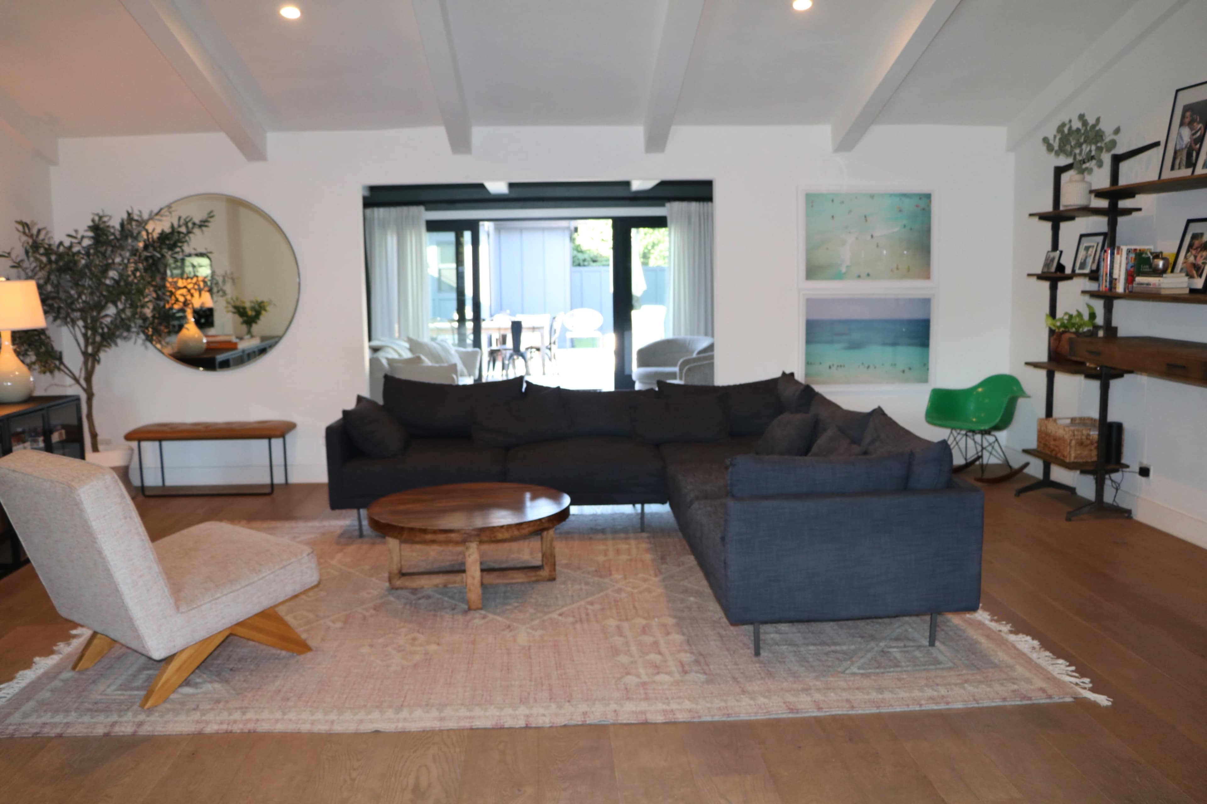 A modern living room features a gray sectional sofa, a round mirror, a wooden coffee table, and two armchairs, with large windows revealing an outdoor space.