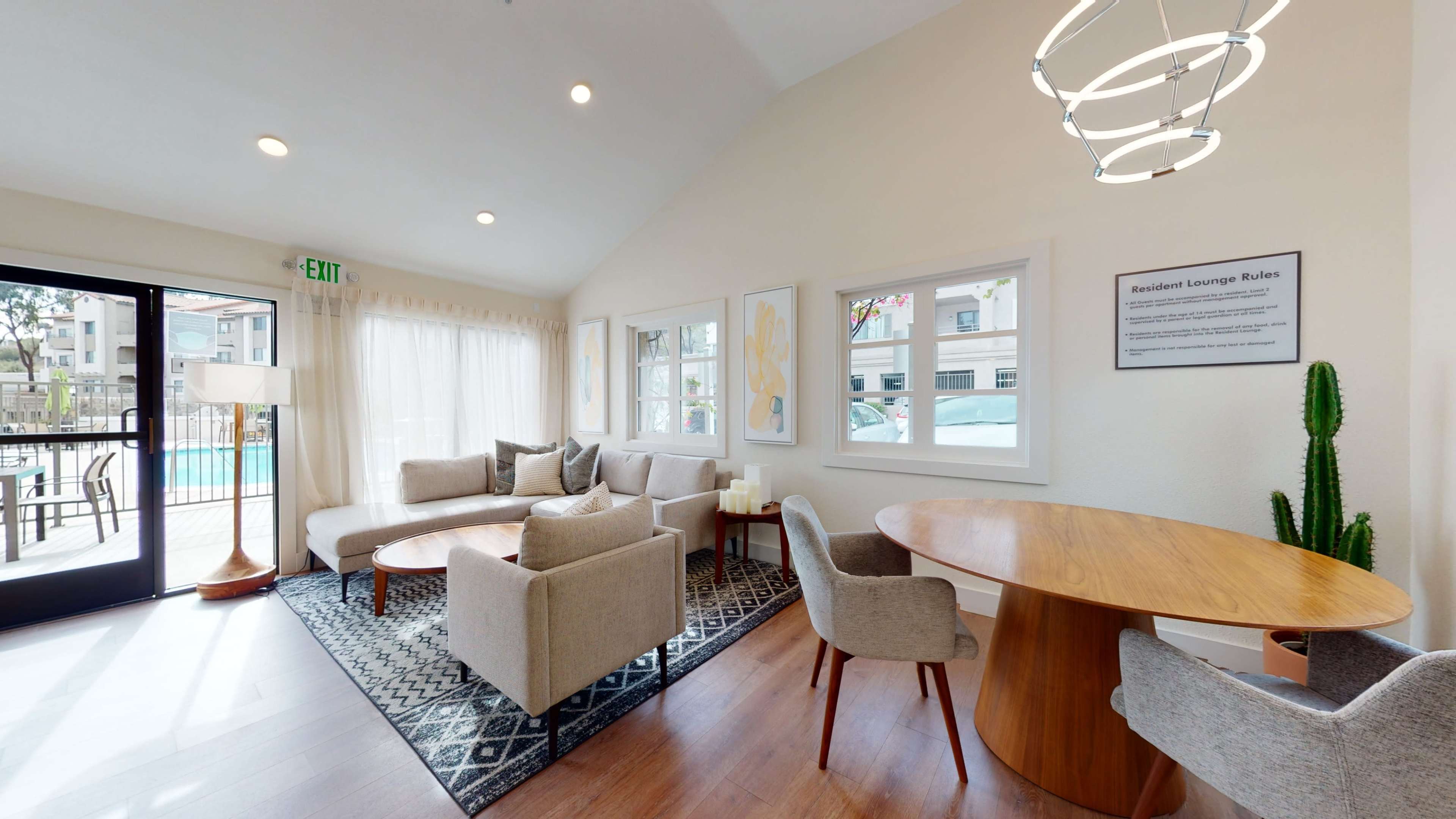 A well-lit lounge area featuring a seating arrangement with grey chairs and a round wooden table, accompanied by large windows overlooking a swimming pool.