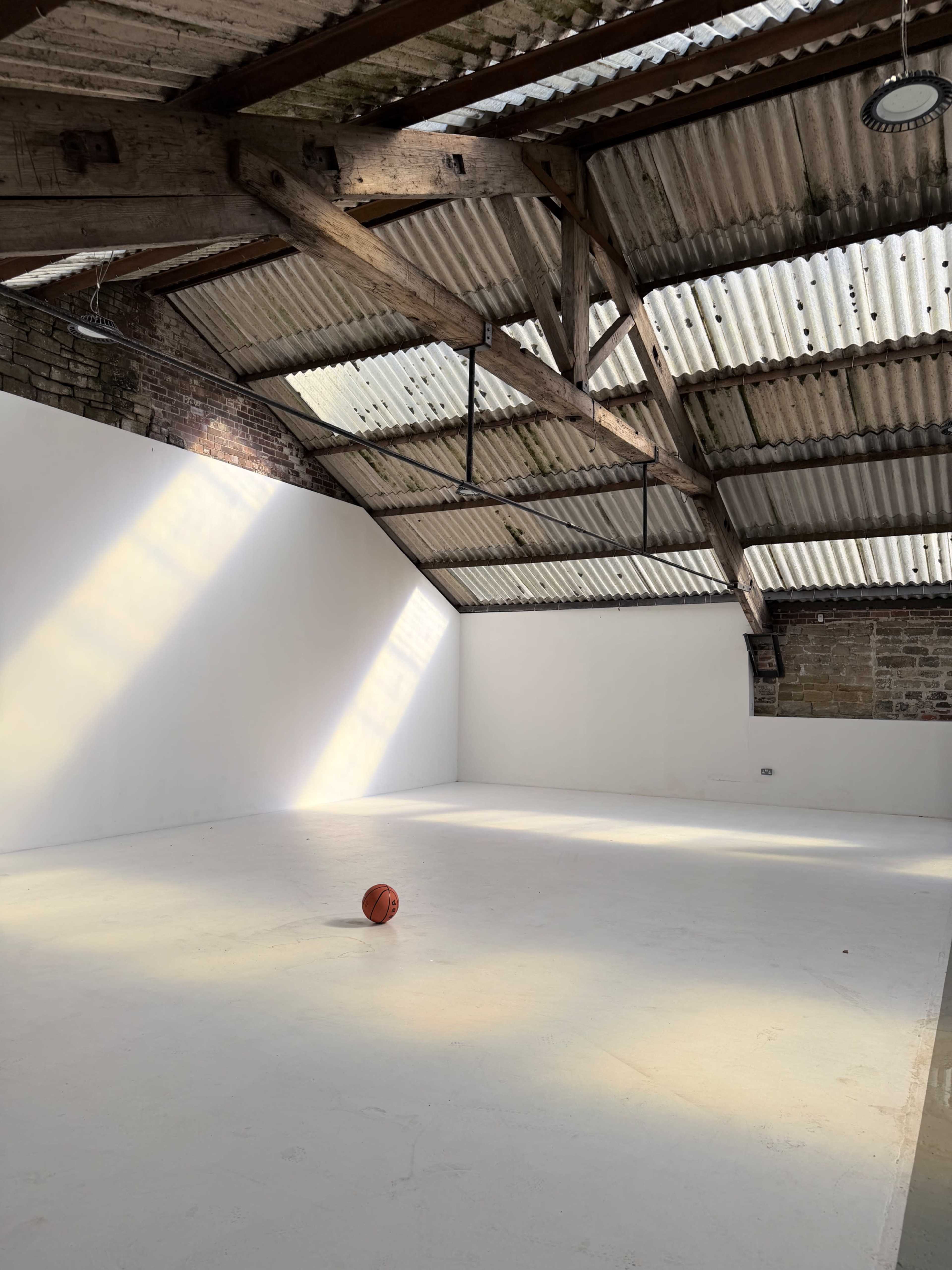 An empty room with a white floor and walls, featuring exposed wooden beams and a single basketball in the center.