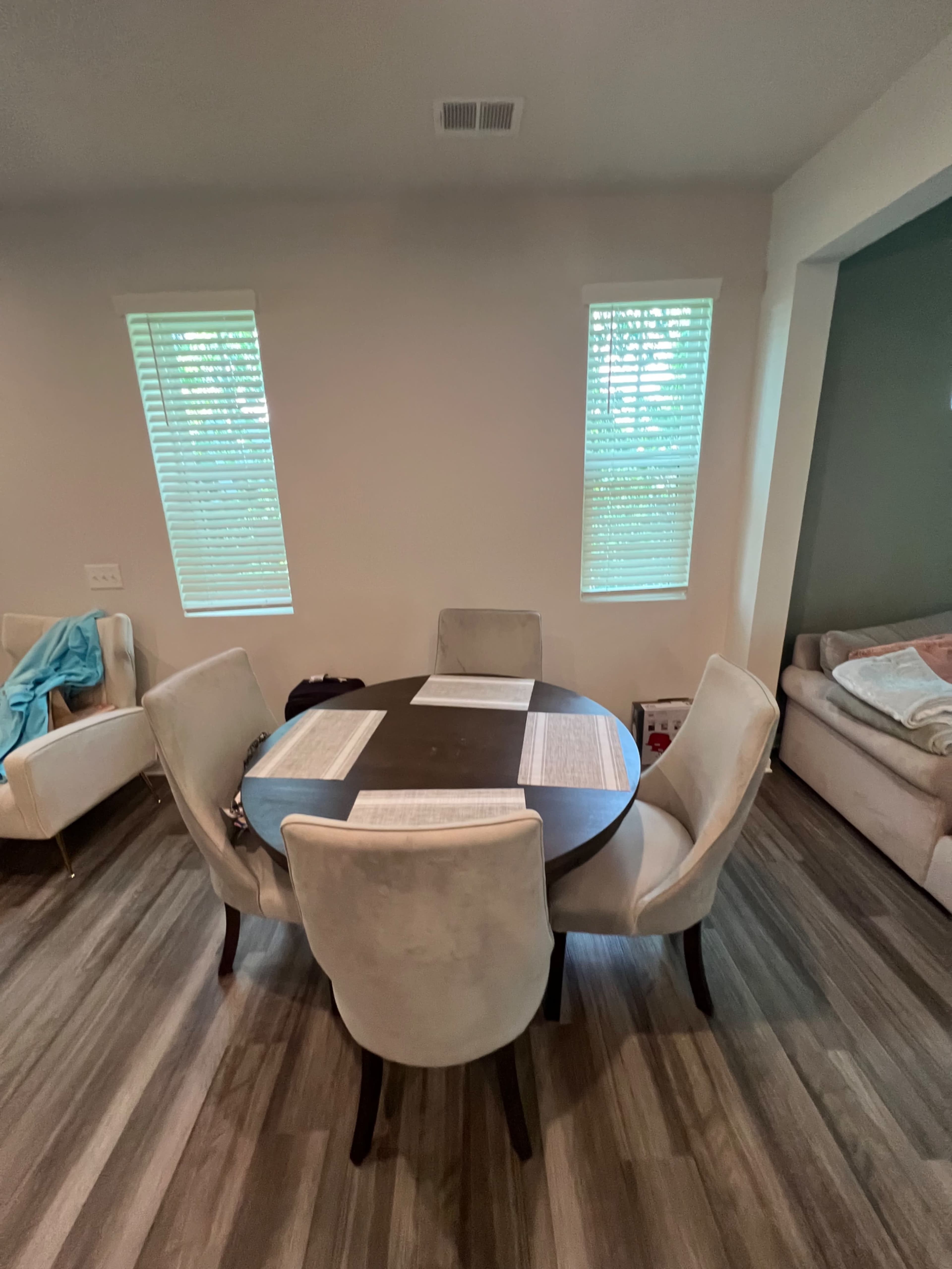 A round dining table with four upholstered chairs is positioned between two windows in a room with wood-like flooring.