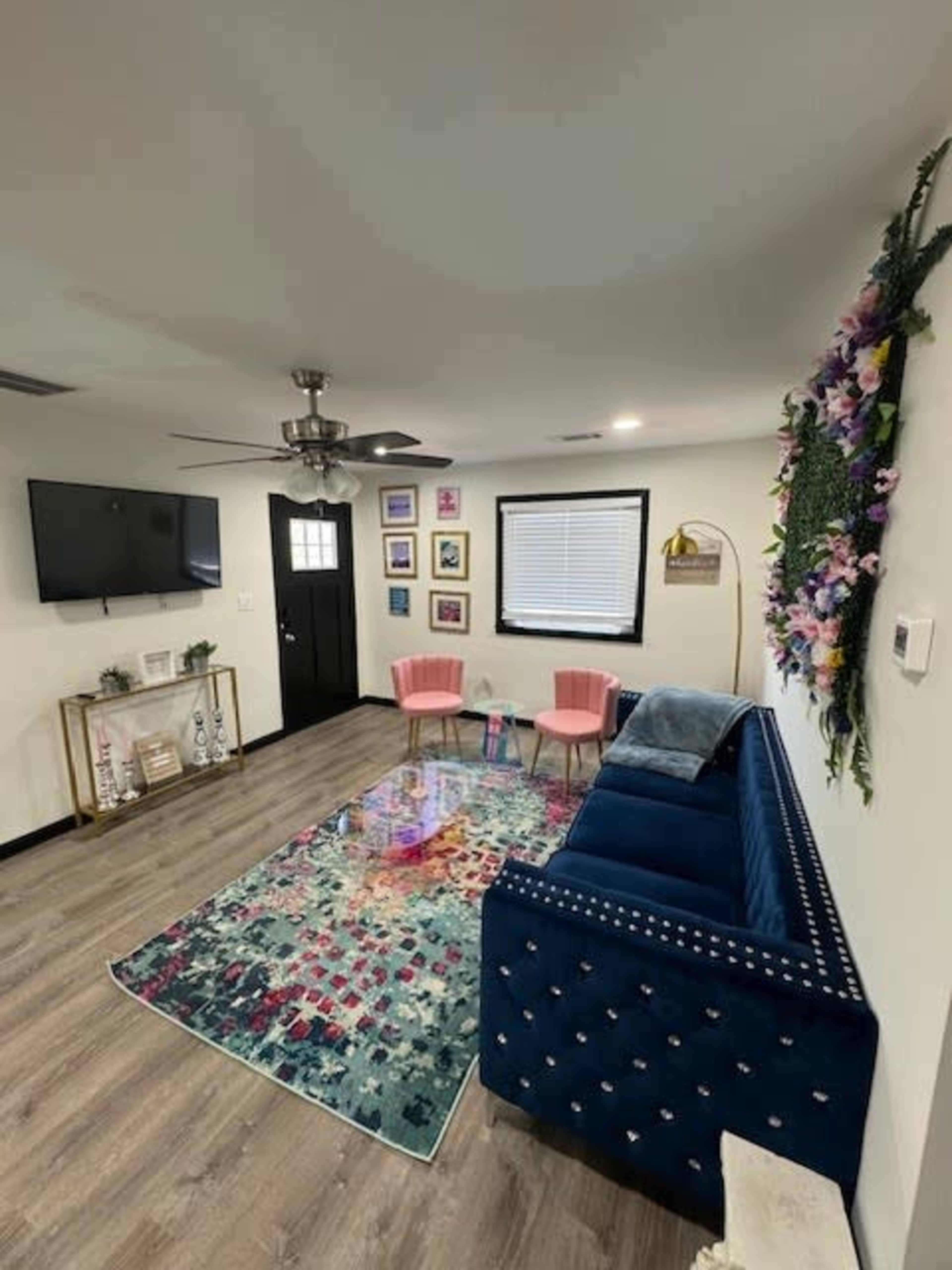 A living room features a blue couch, two pink chairs, a colorful area rug, a television mounted on the wall, and a door leading outside.