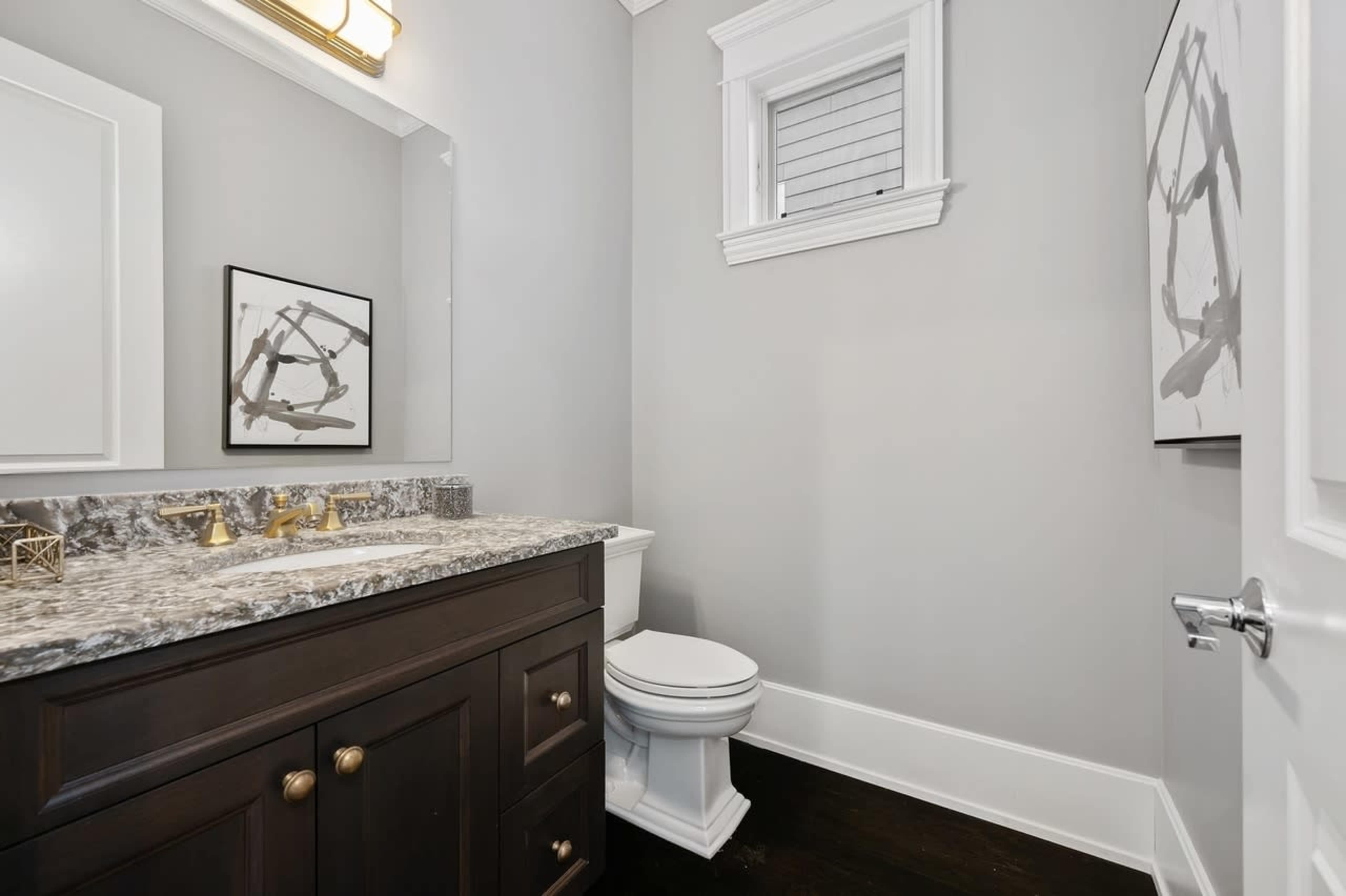 A modern bathroom featuring a dark wood vanity with a granite countertop, a white toilet, and framed wall art against a light gray wall.
