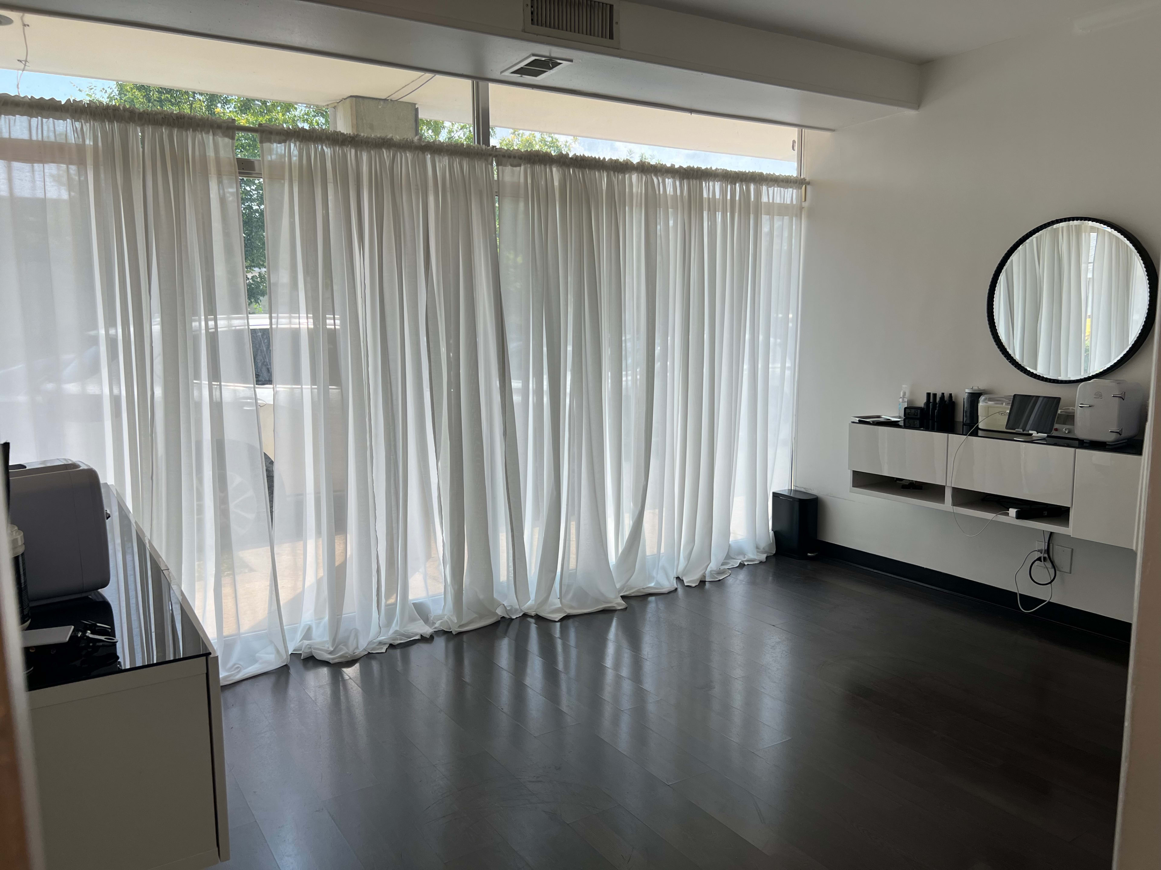 A spacious room with white sheer curtains covering large windows, a round mirror on the wall, and minimal furniture including a low shelf and appliances.