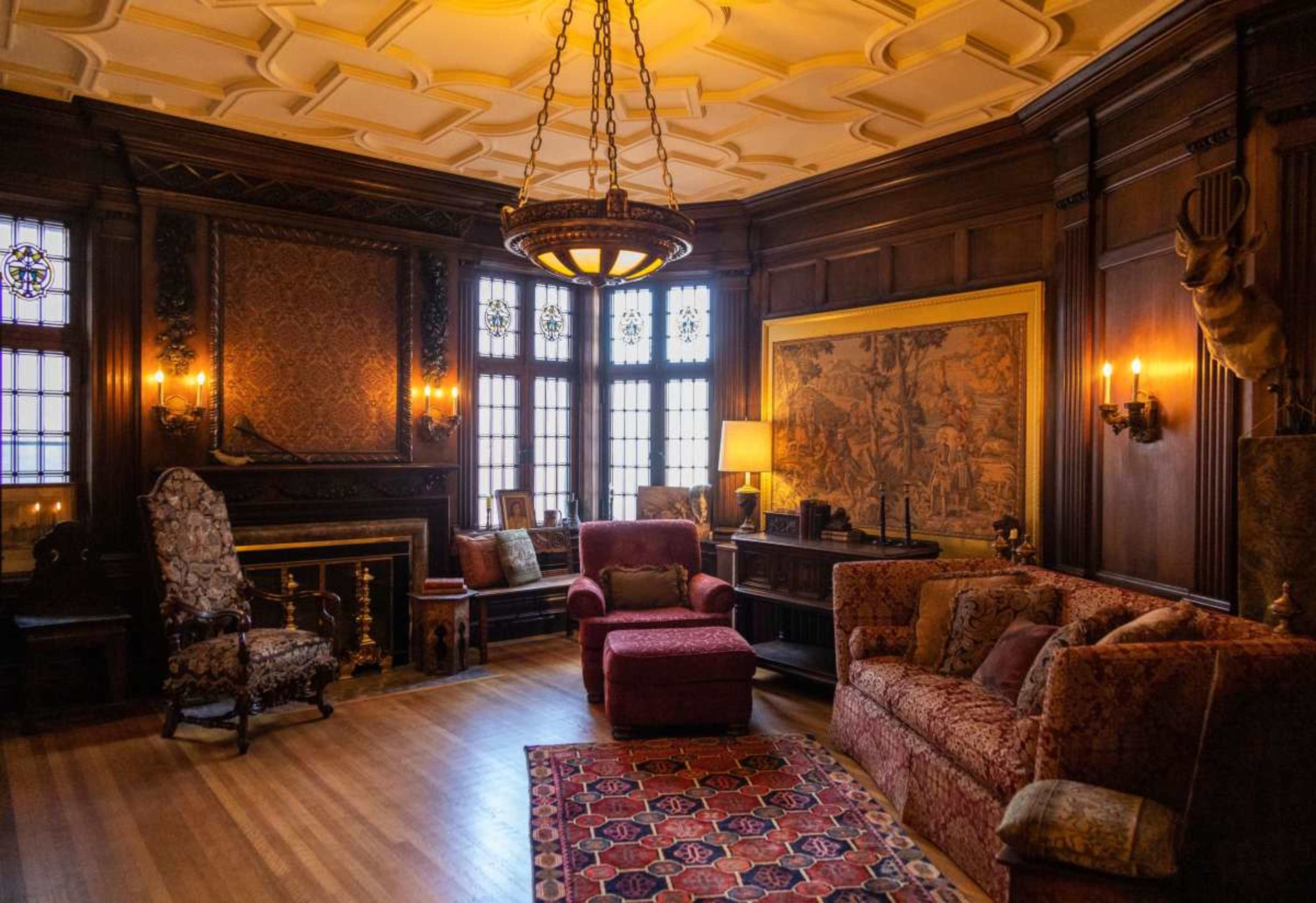 The room features dark wooden paneling, ornate furniture, and a large tapestry, with light streaming through decorative windows.
