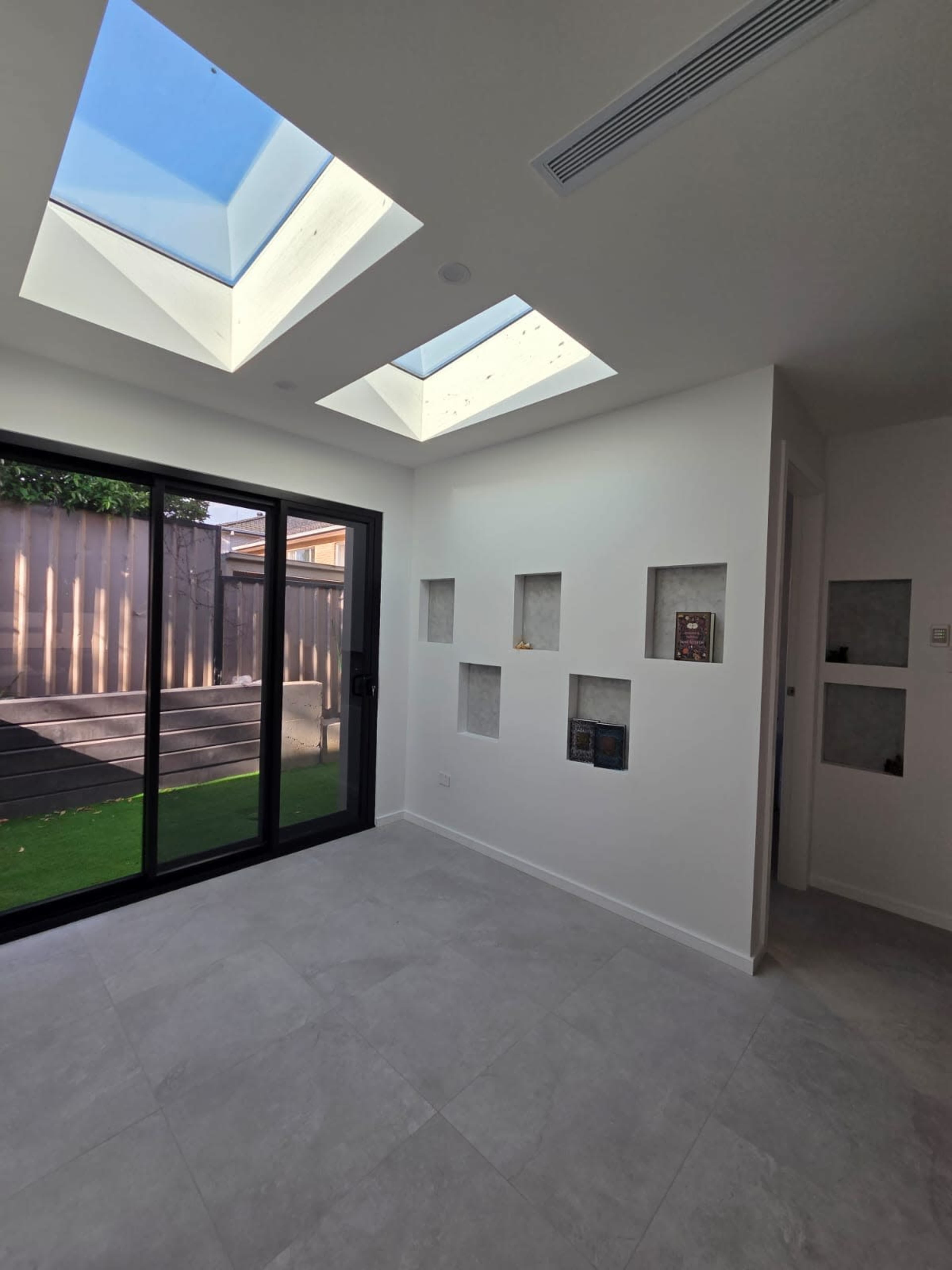 The image shows a bright, minimalist living space with large skylights and a sliding glass door leading to a patio.