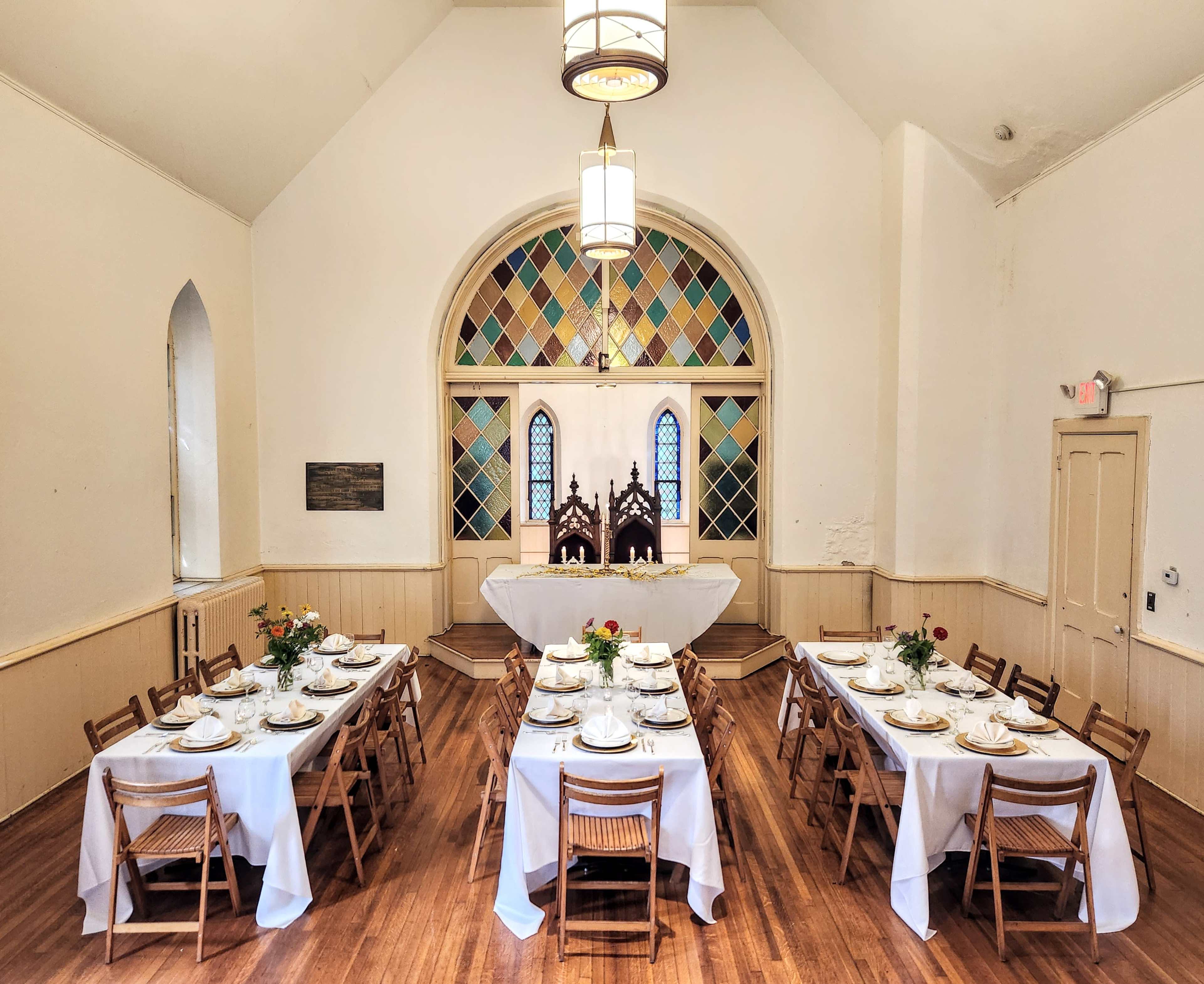 A dining hall features two long tables set with white tablecloths and plates, positioned beneath stained glass windows.