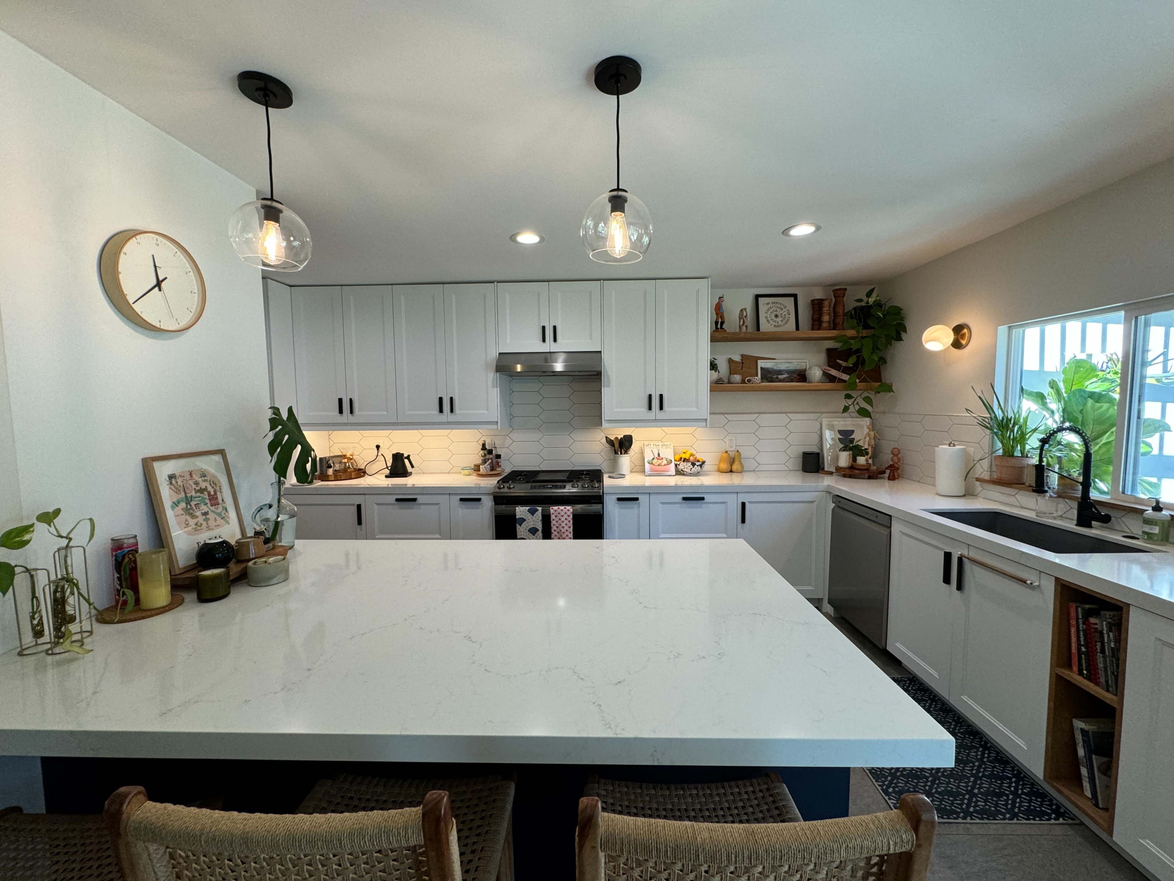 A modern kitchen features a large white countertop, sleek cabinetry, and a window with greenery, illuminated by pendant lights.