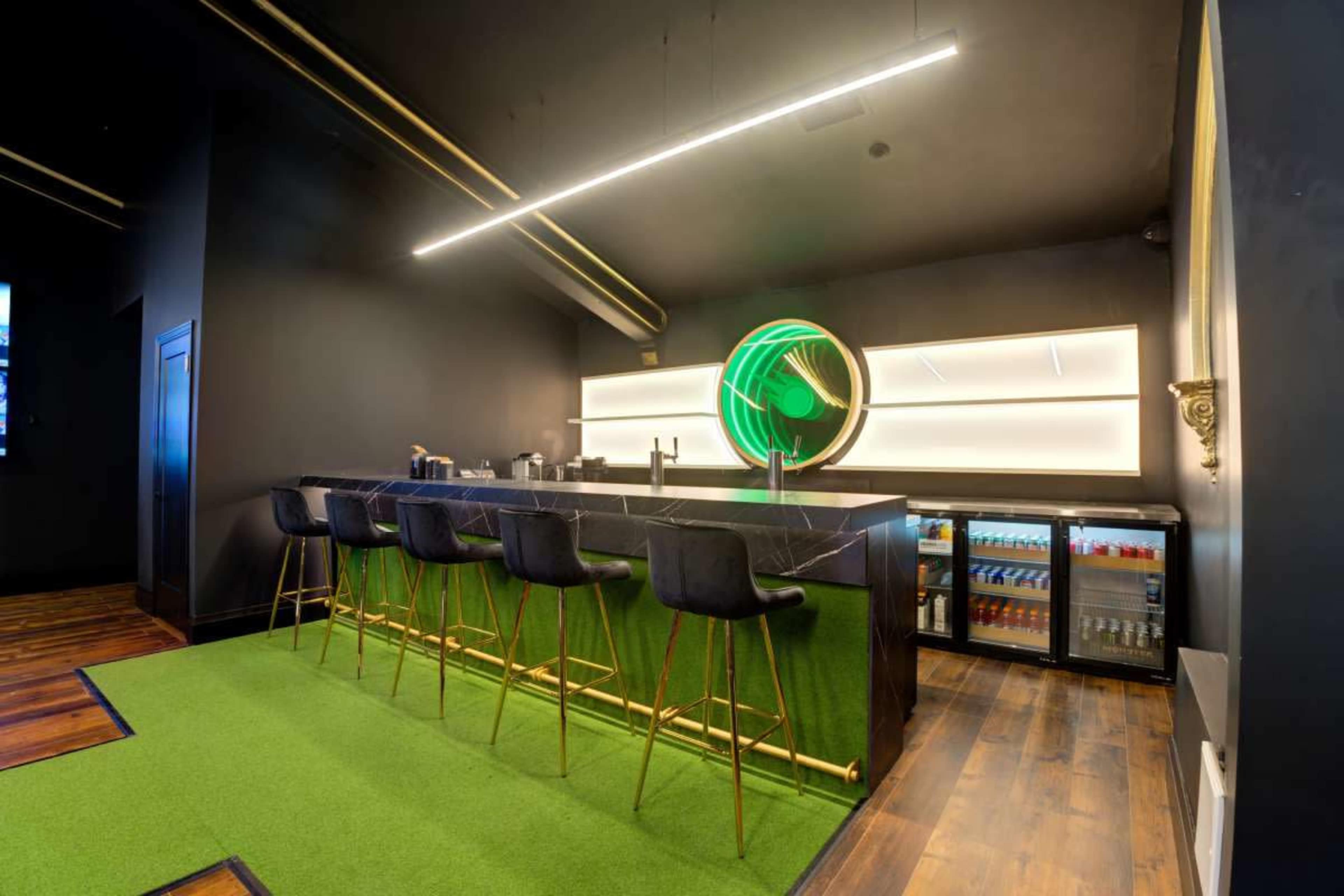 The image shows a modern bar with a black interior, featuring green barstools, a marble countertop, and a circular neon decoration.