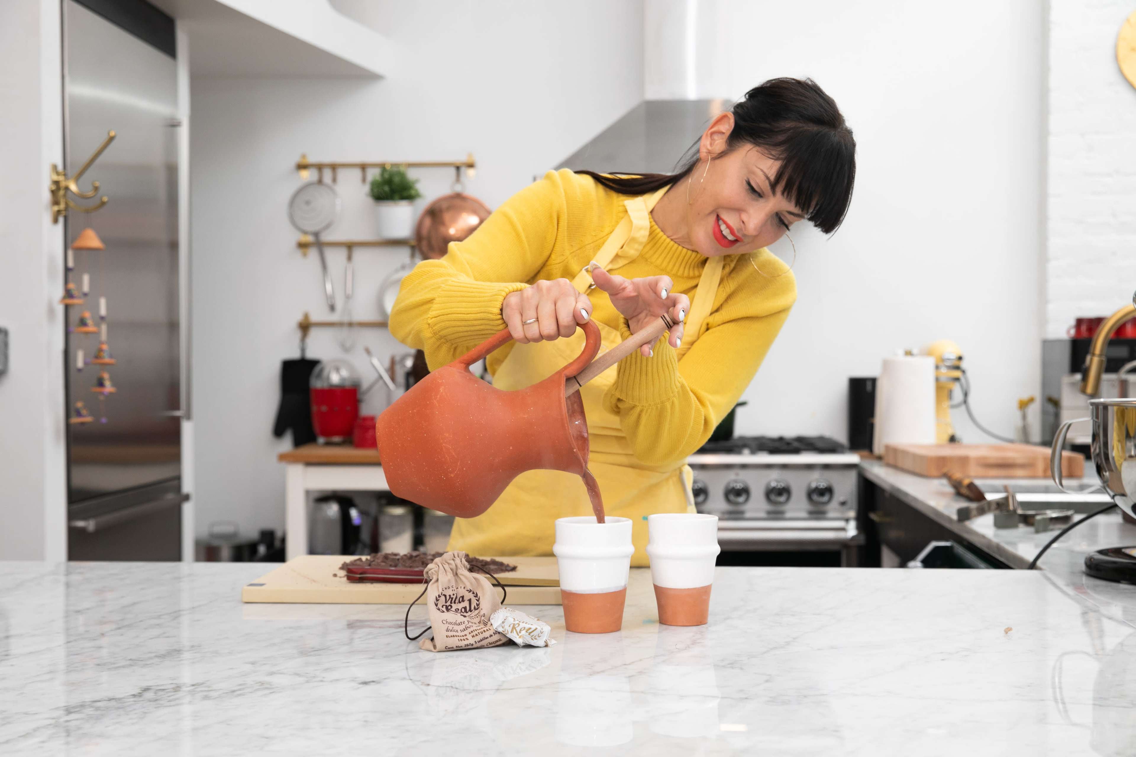 A woman in a yellow sweater is pouring a beverage from a clay pitcher into two white cups on a marble kitchen countertop.