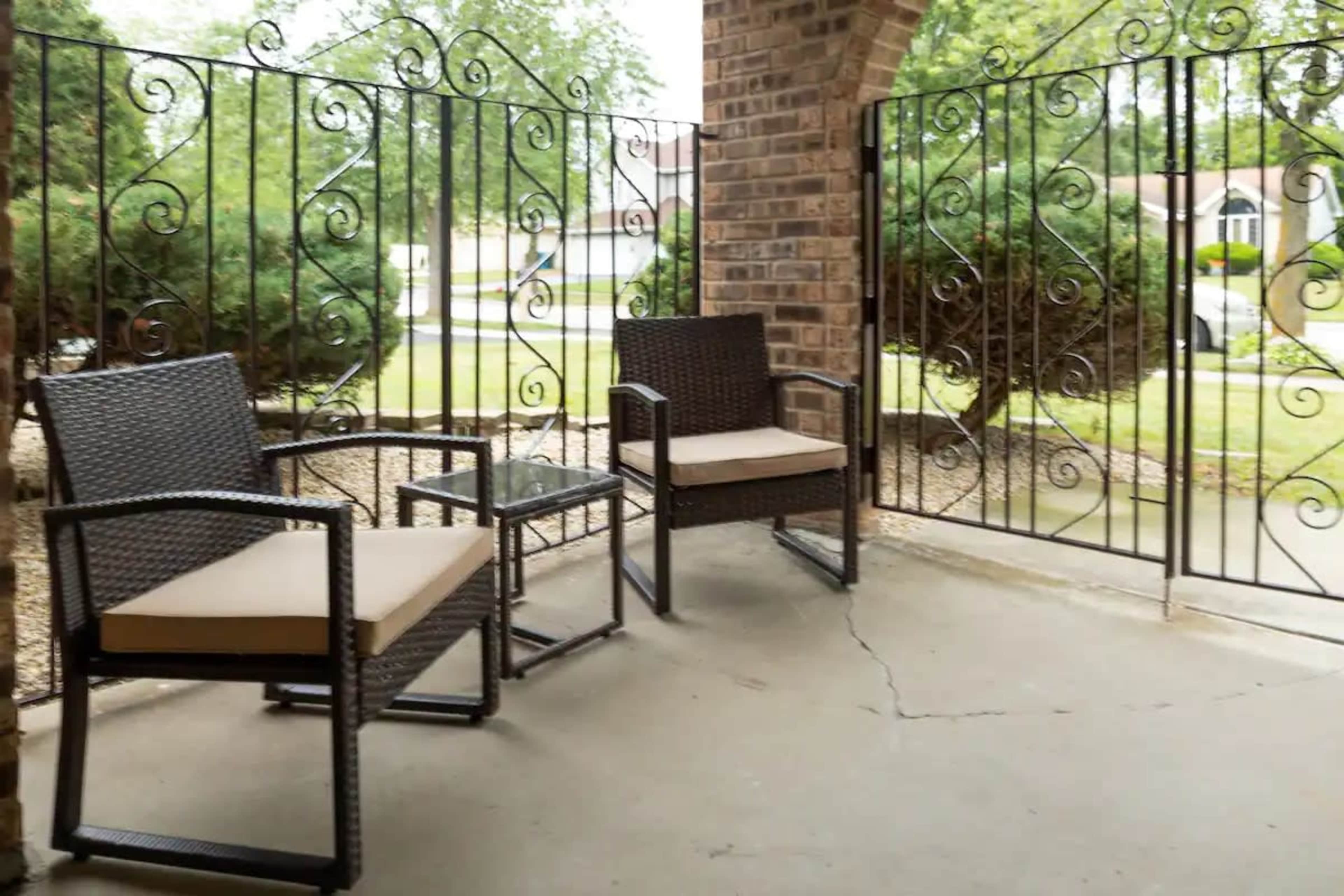 The image shows a small porch area with two wicker chairs, a glass-topped table, and decorative wrought iron fencing.